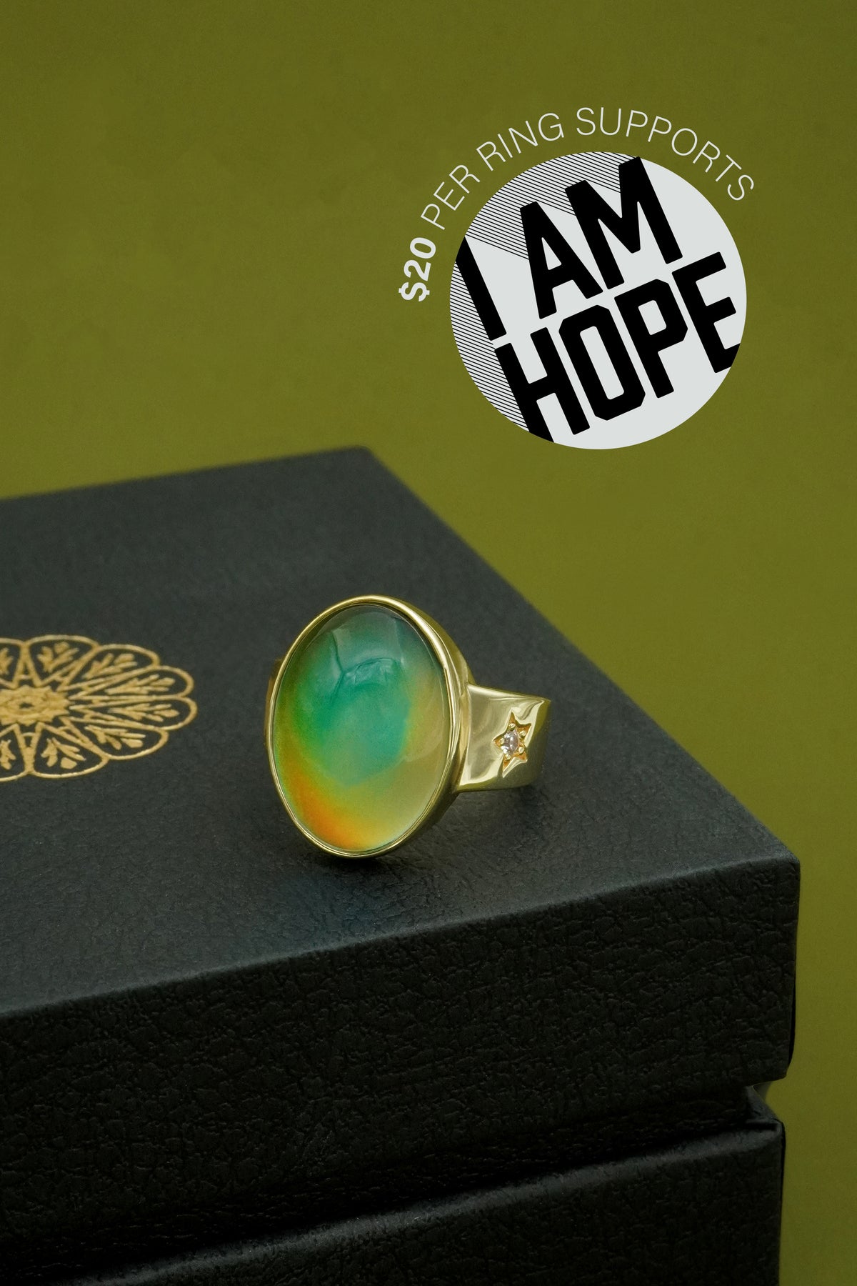 Ring with green and gold stone on a black box against a green background, featuring 'I AM HOPE' logo.