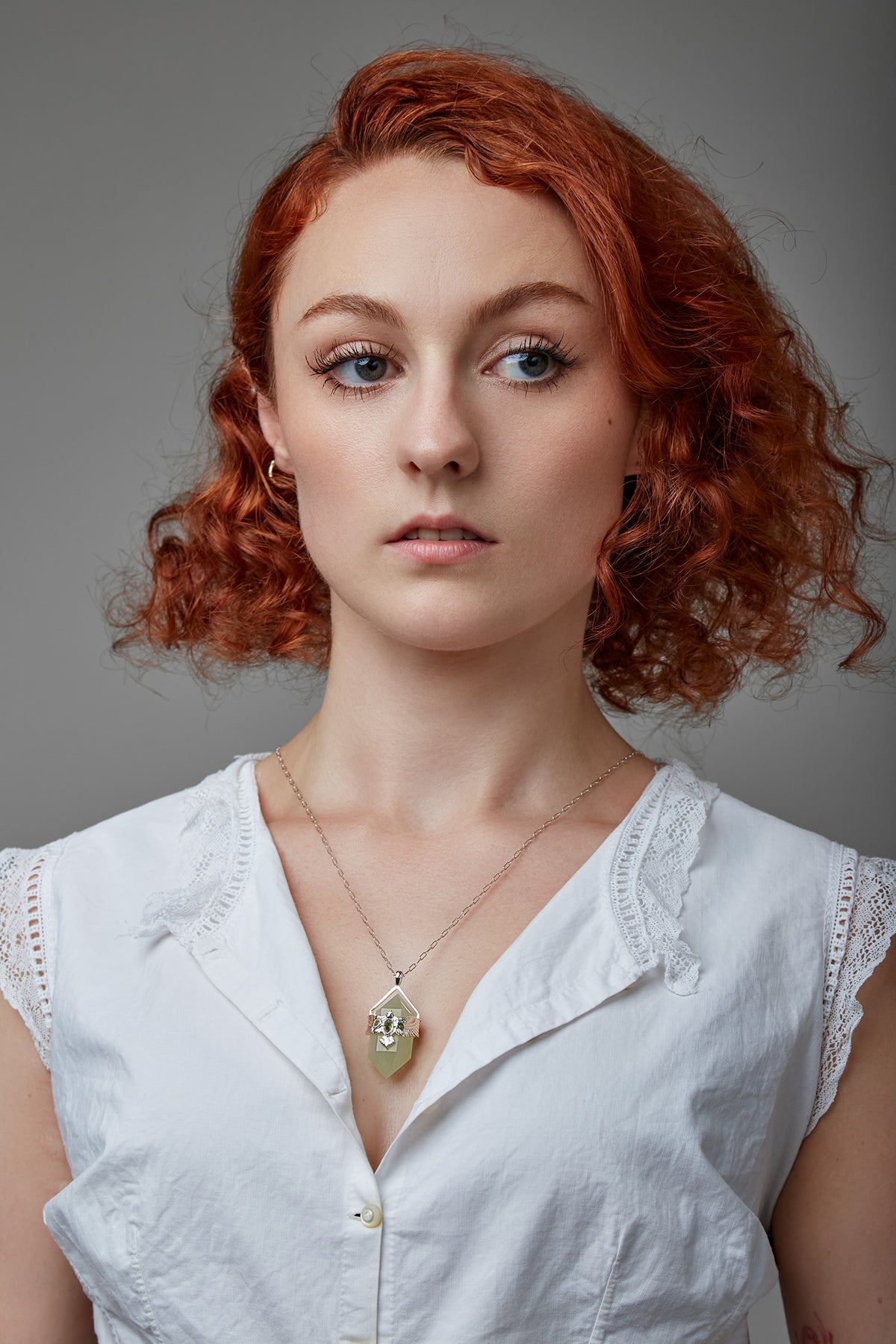 Woman with red hair wearing a white blouse against a gray background wearing green crystal necklace