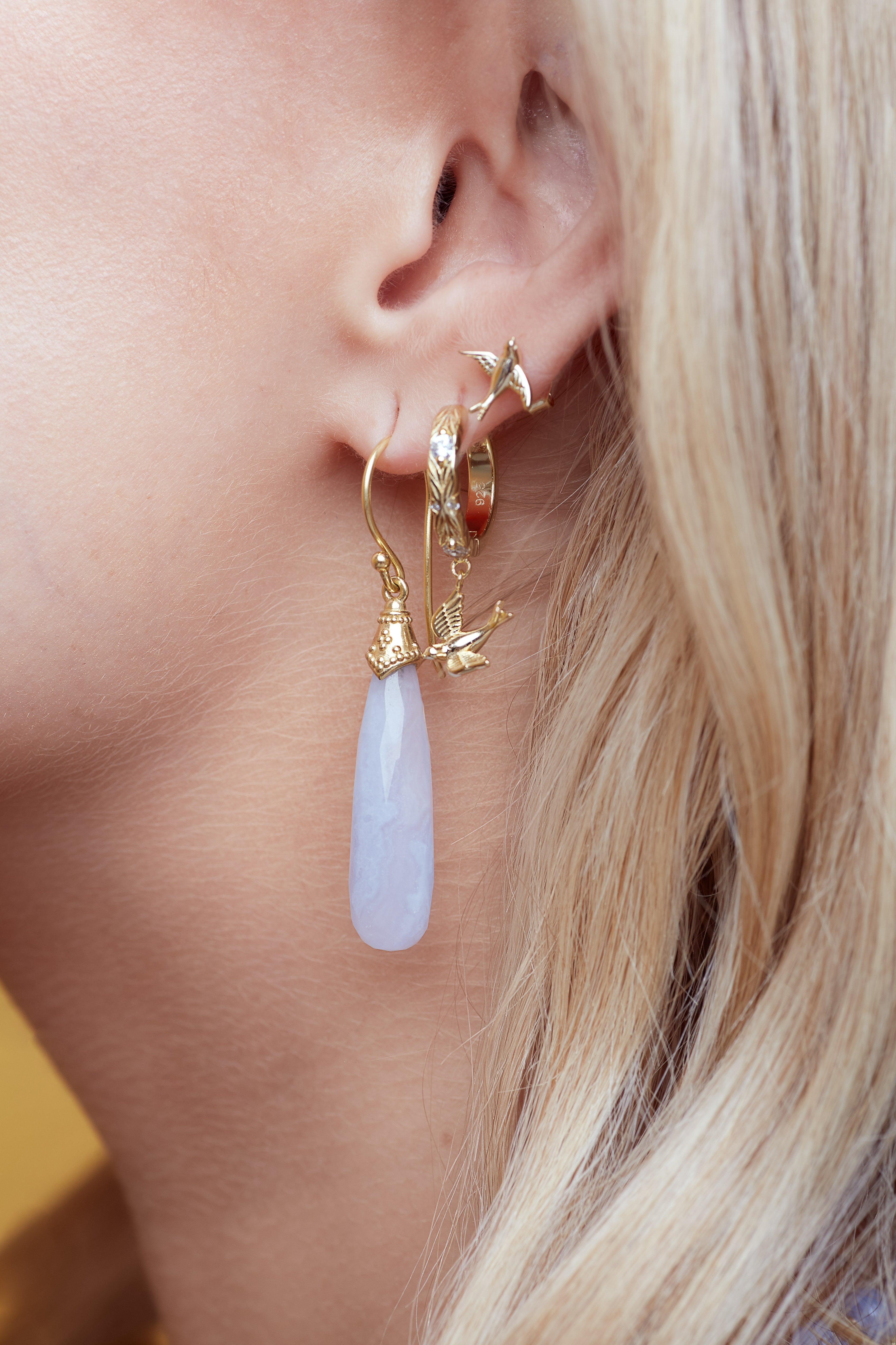 Close-up of a person wearing gold hoop earrings with a teardrop-shaped pearl-like pendant.