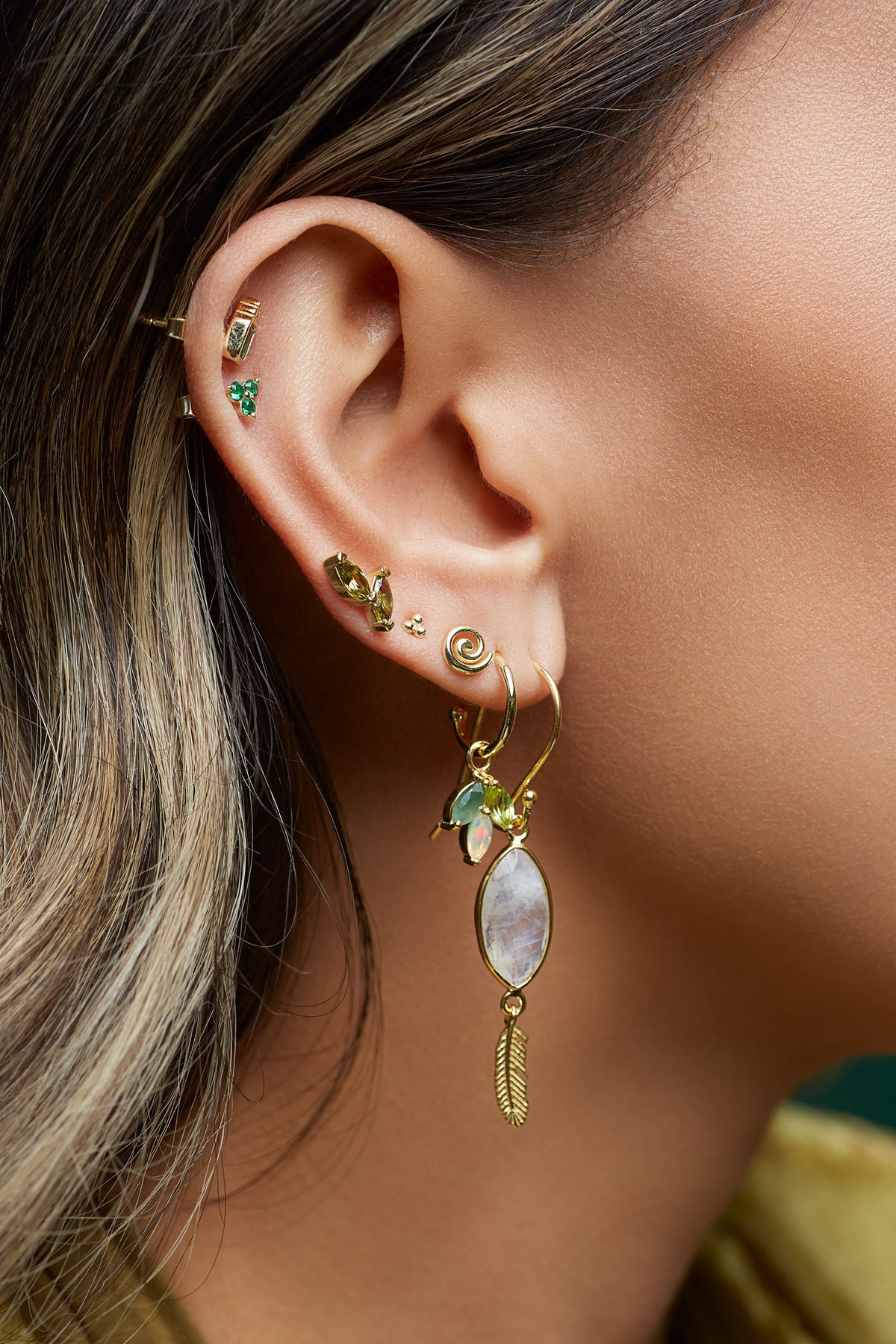 Close-up of an ear with gold earrings and green gemstones.