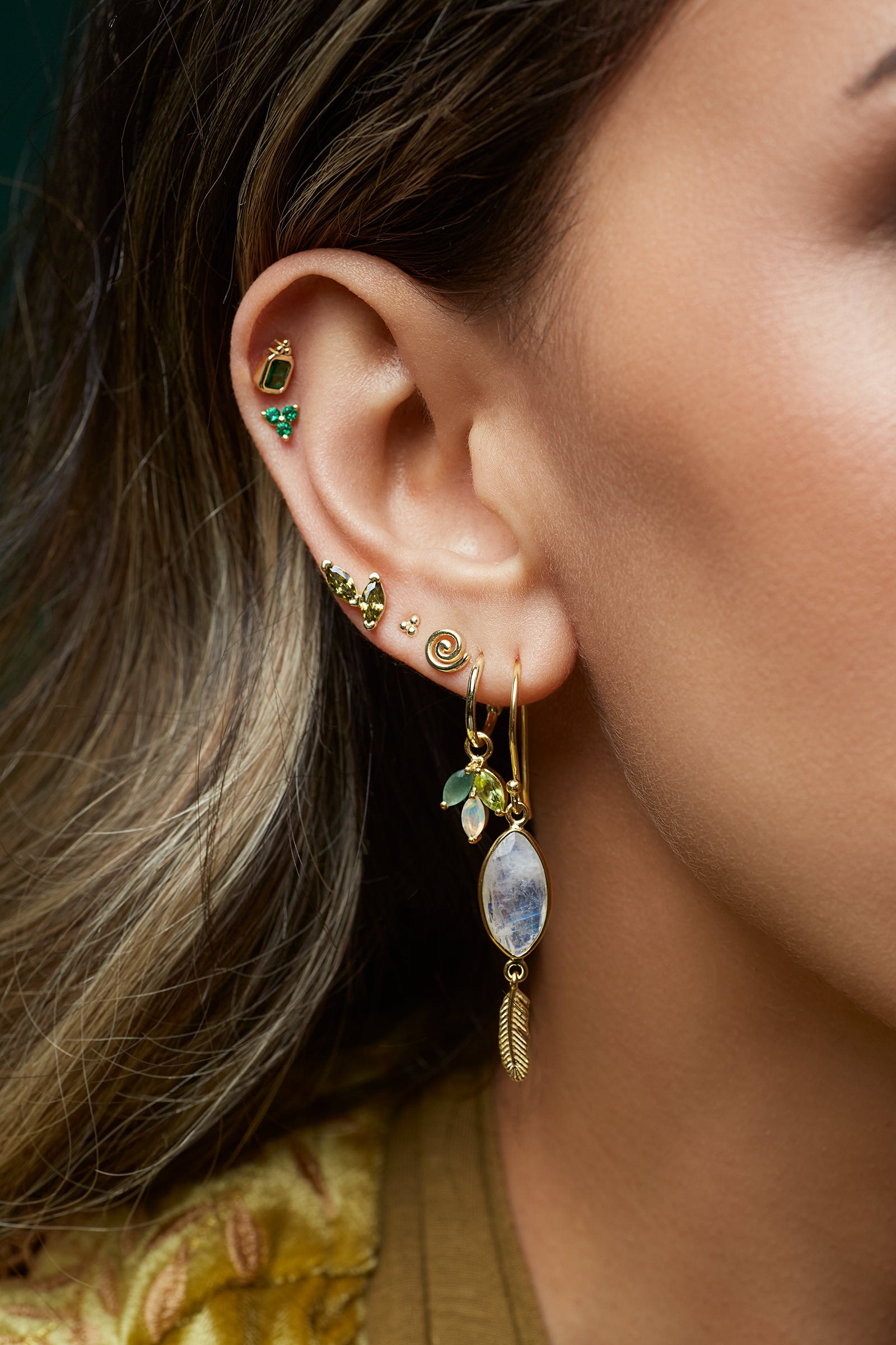 Close-up of an ear with gold earrings featuring green stones and a large dangling earring.