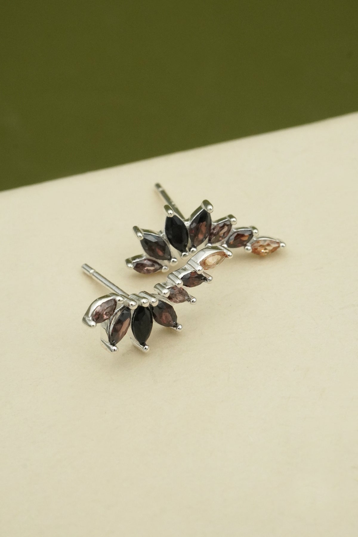 Silver leaf-shaped earrings with gemstones on a beige surface and green background