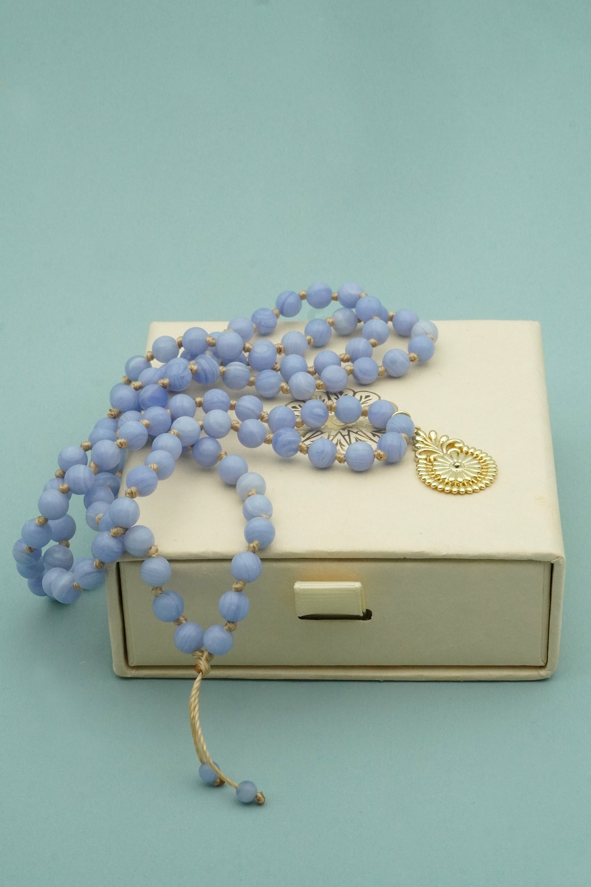 Blue beaded necklace with a gold charm on a beige box against a light blue background
