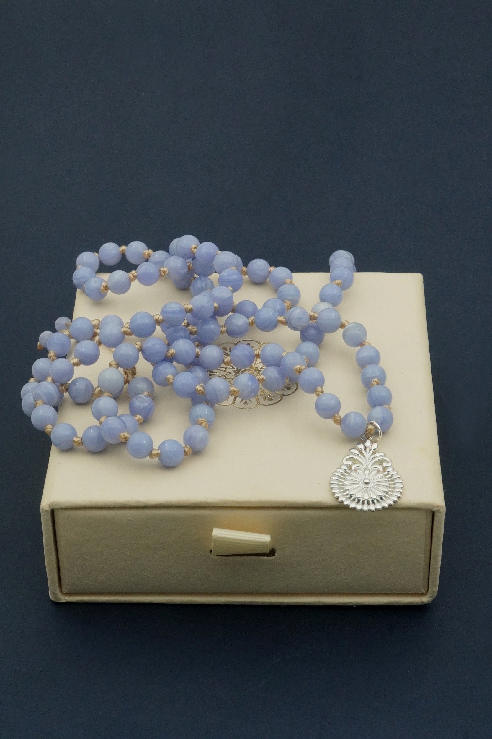 Blue beaded necklace with a silver pendant on a beige box against a dark background