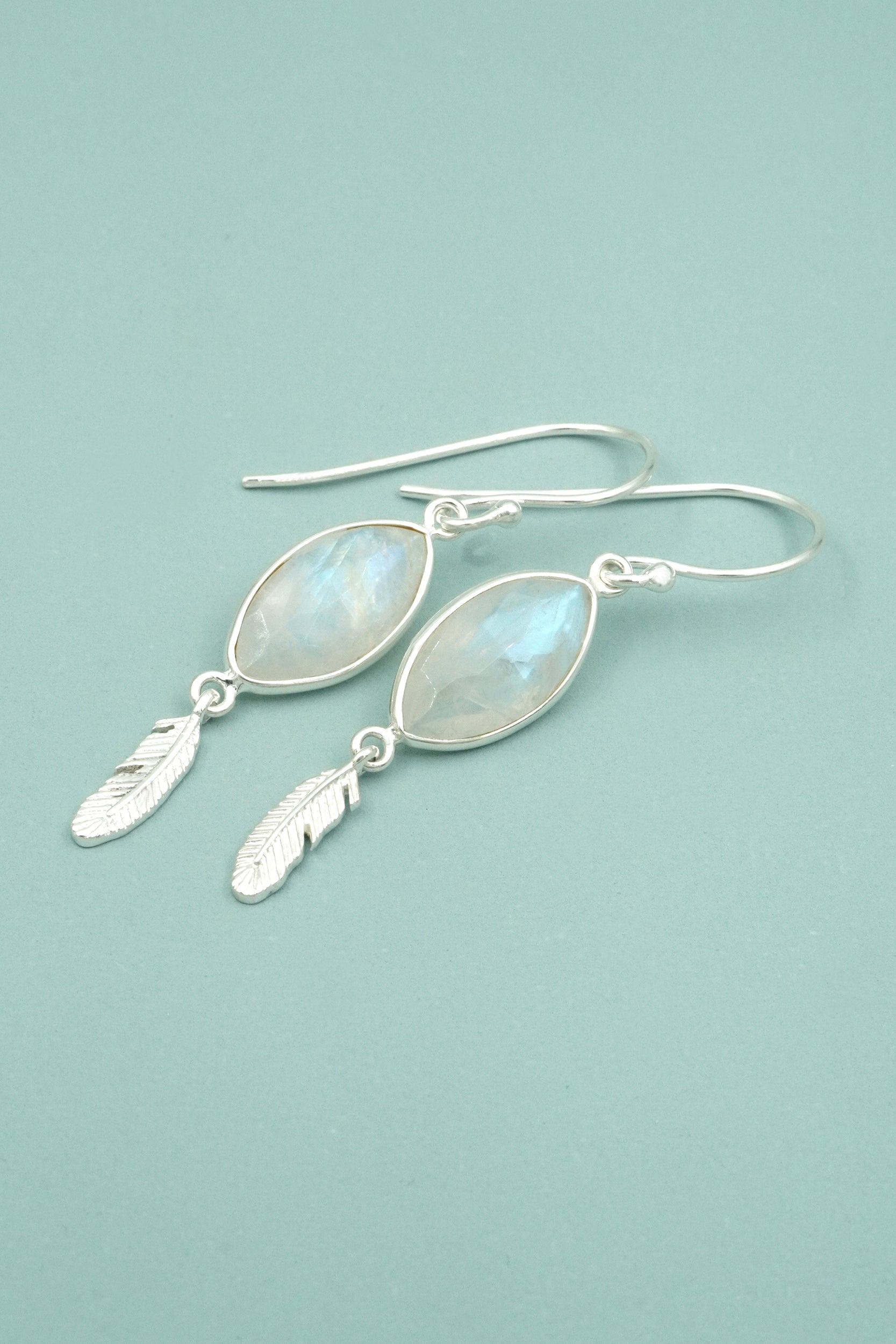 Silver earrings with moonstone stones and feather charms on a light blue background
