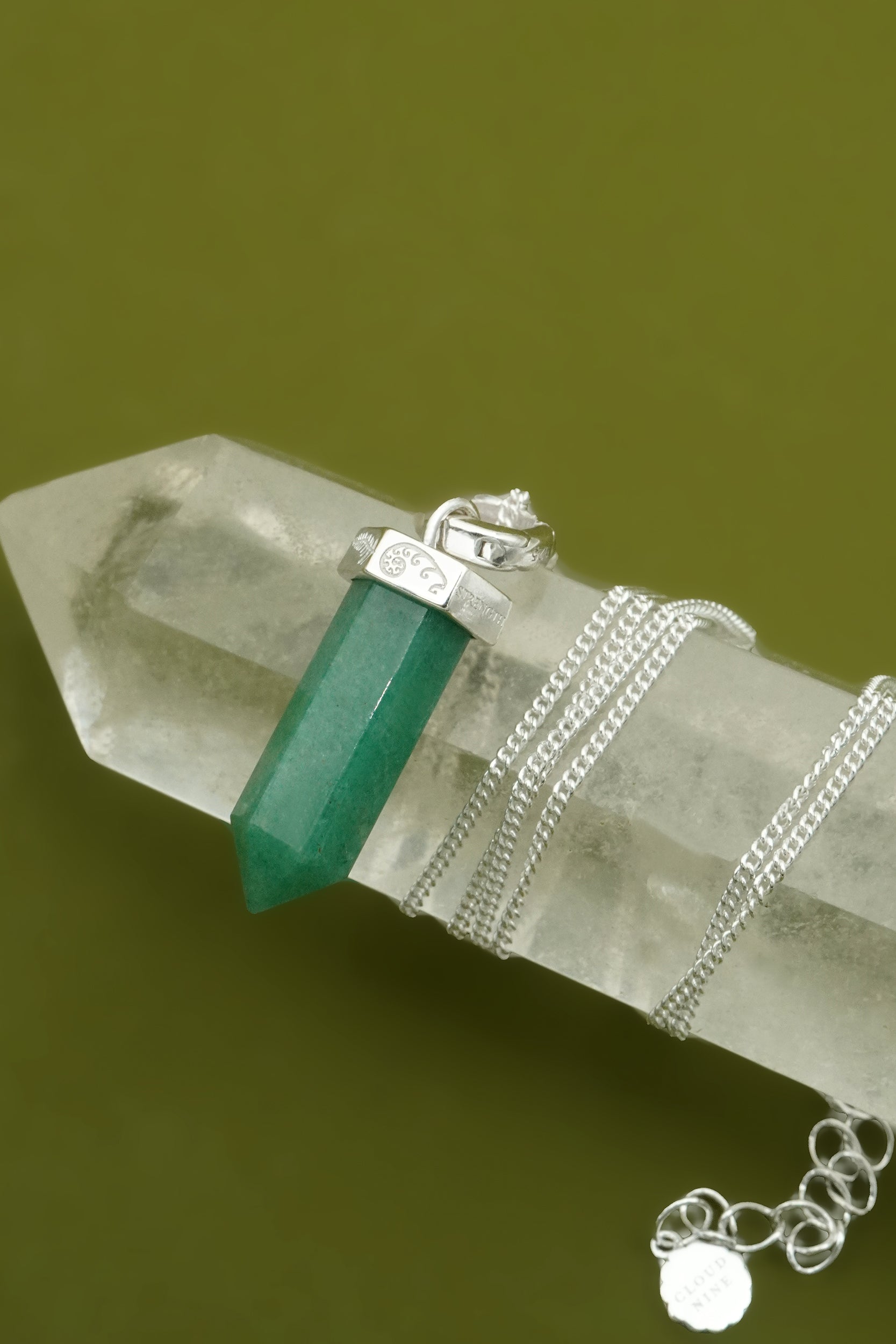 Green crystal pendant on a silver chain with a clear crystal base against a green background featuring fern and koru detailing.