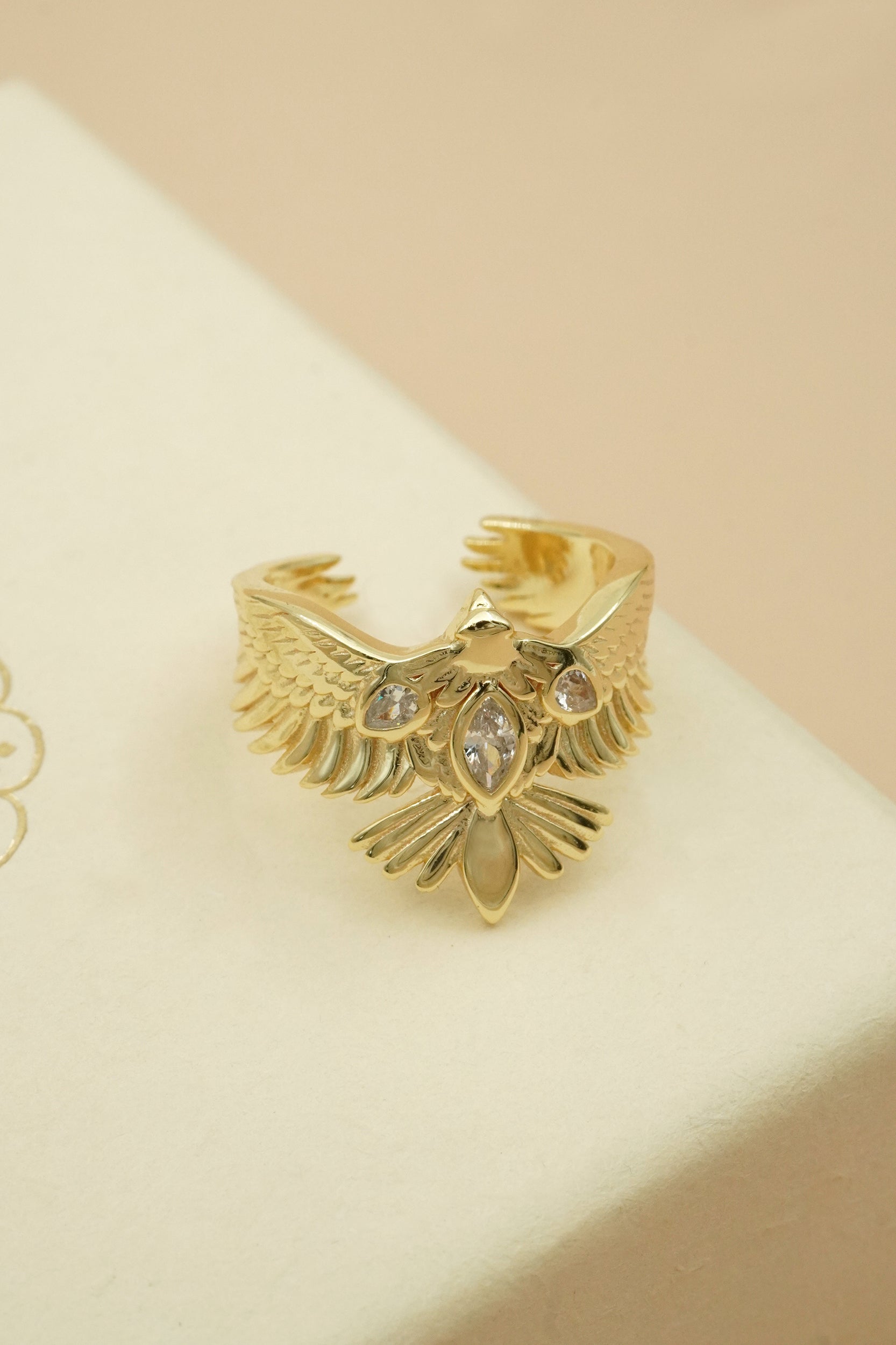 Gold phoenix hawk bird ring with gemstones on a beige background