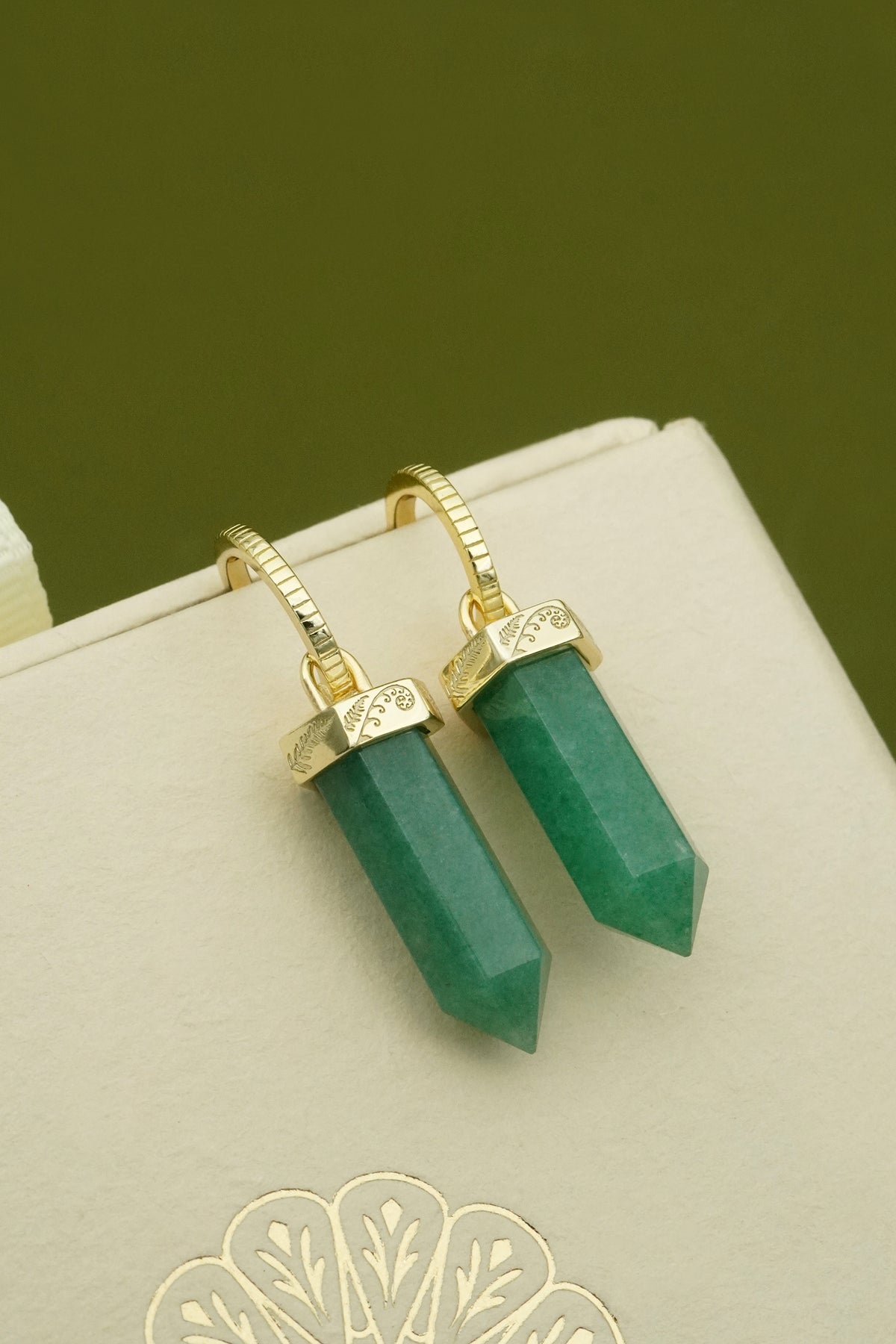 Green aventurine grounding crystal earrings with gold koru and fern detailing on a beige background
