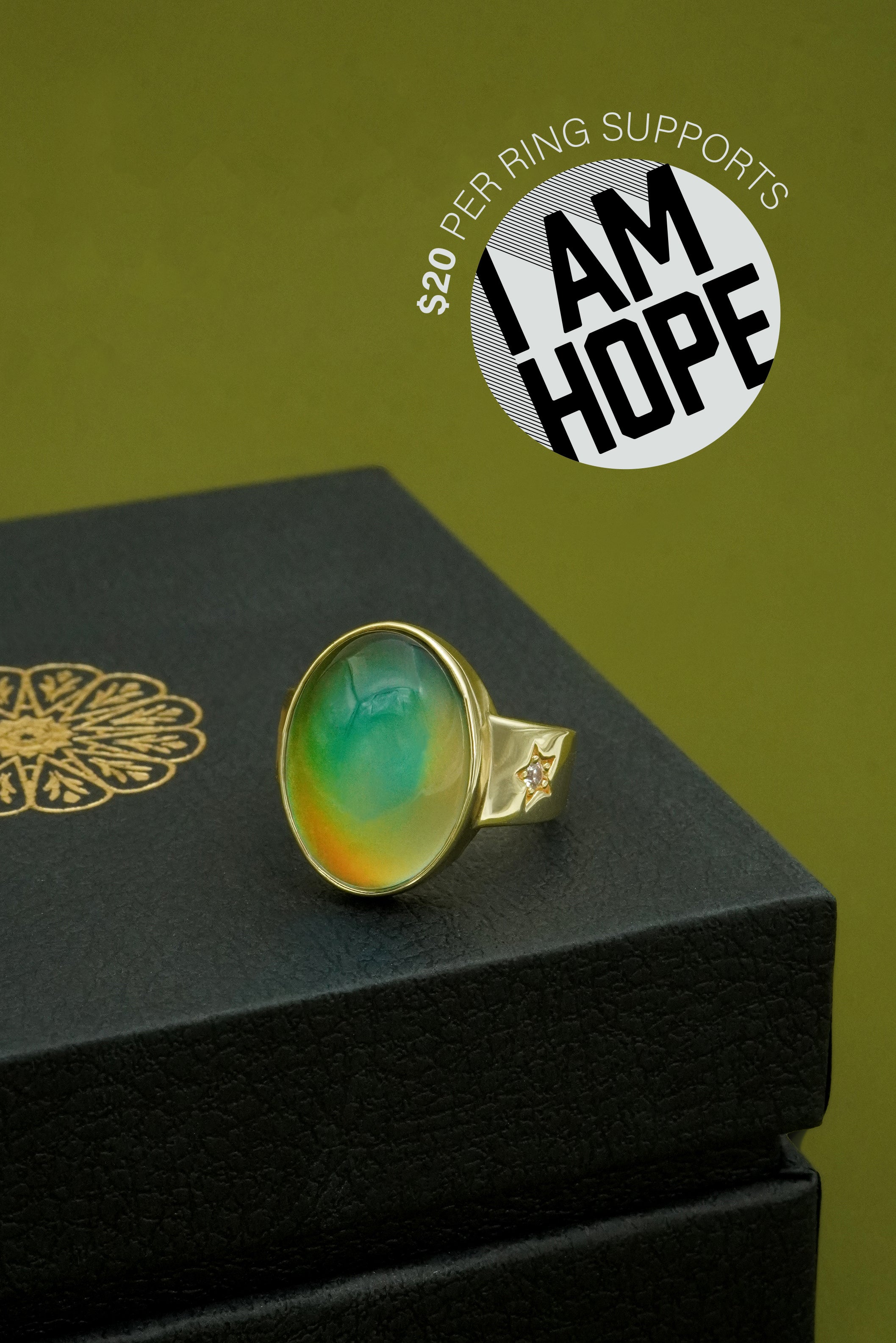 Ring with green and gold stone on a black box against a green background, featuring 'I AM HOPE' logo.