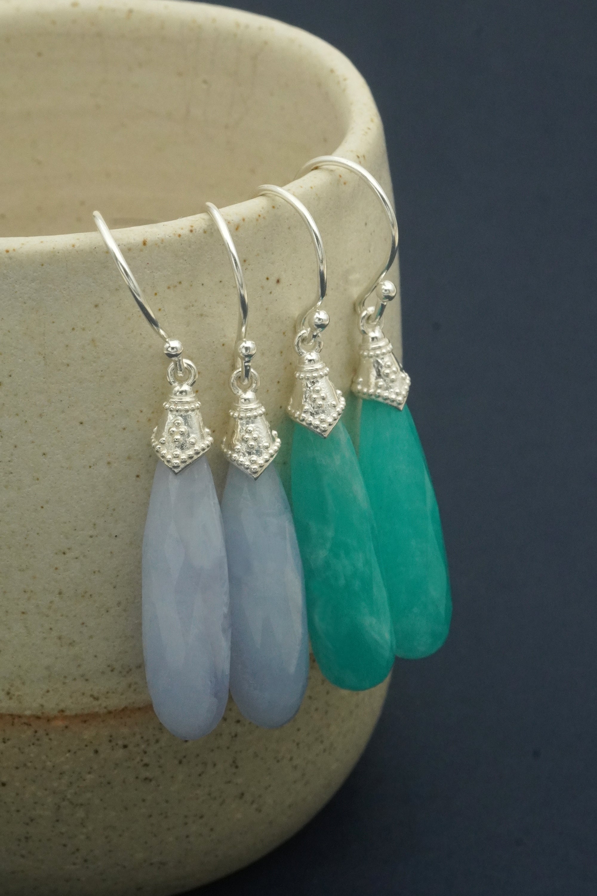 Three crystal teardrop-shaped earrings in teal, light blue, and clear hanging on a hook against a dark background.