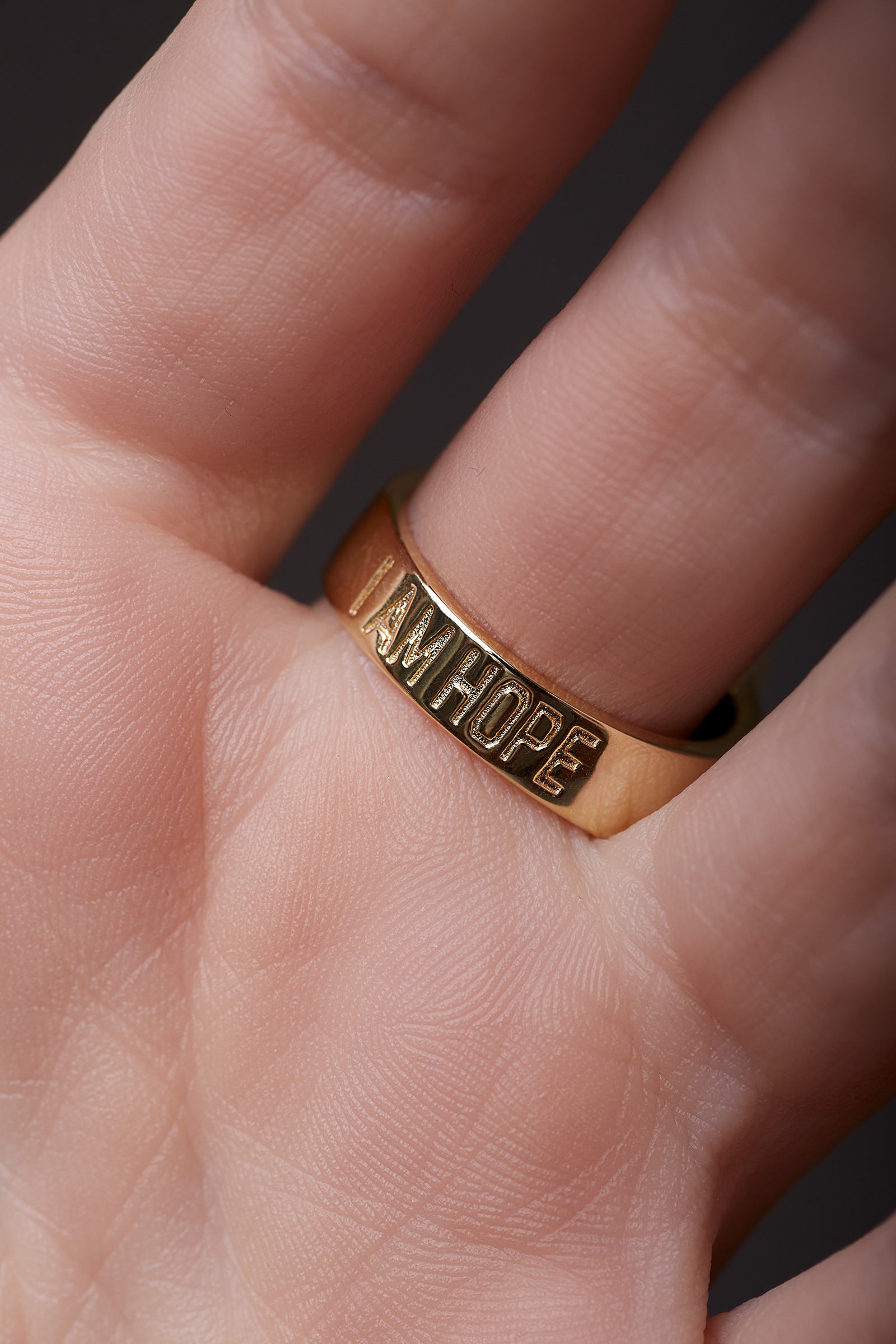 Gold ring with 'I AM HOPE' engraving on a finger against a dark background