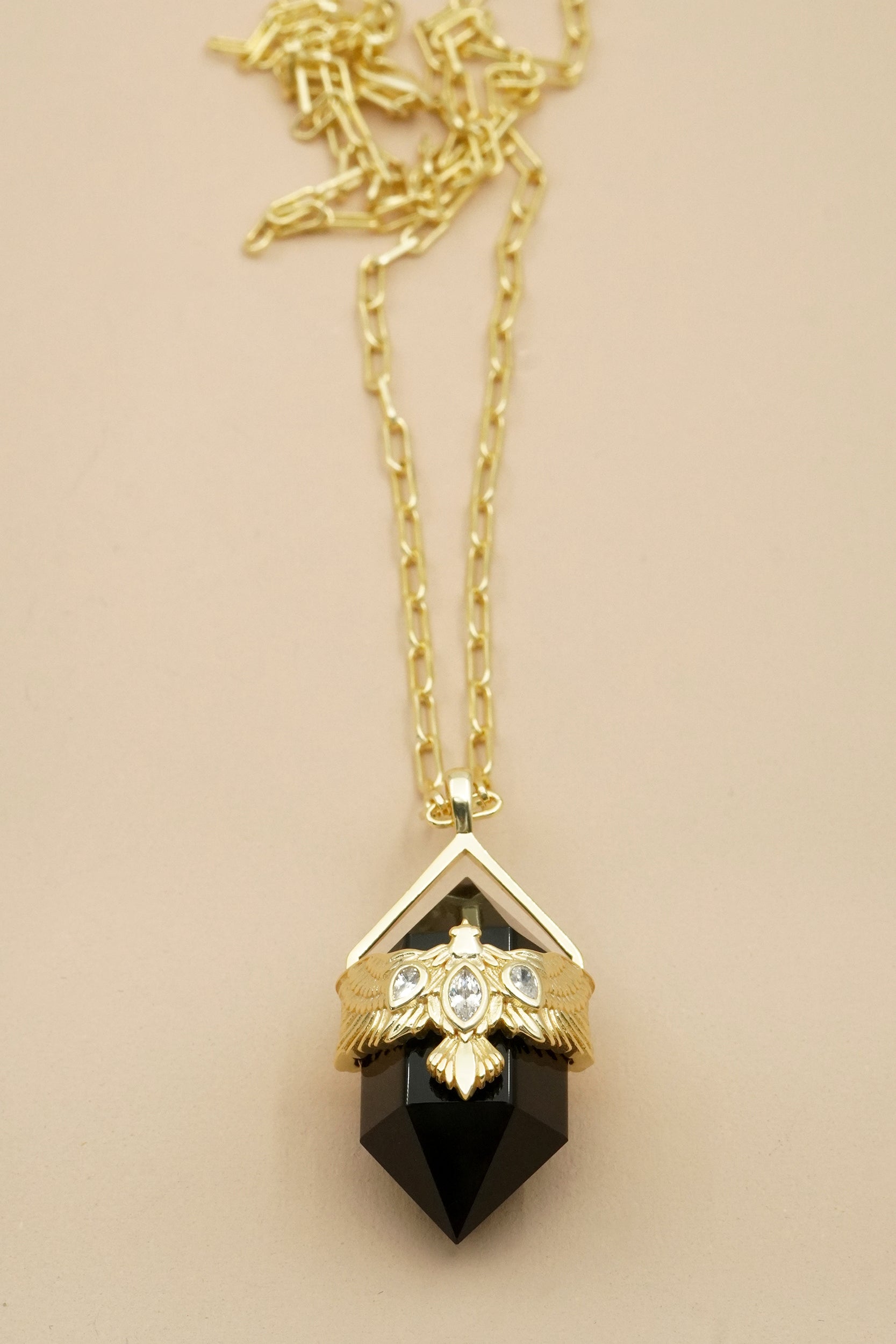 Gold Hawk soar necklace with a protective black agate gemstone pendant on a beige background
