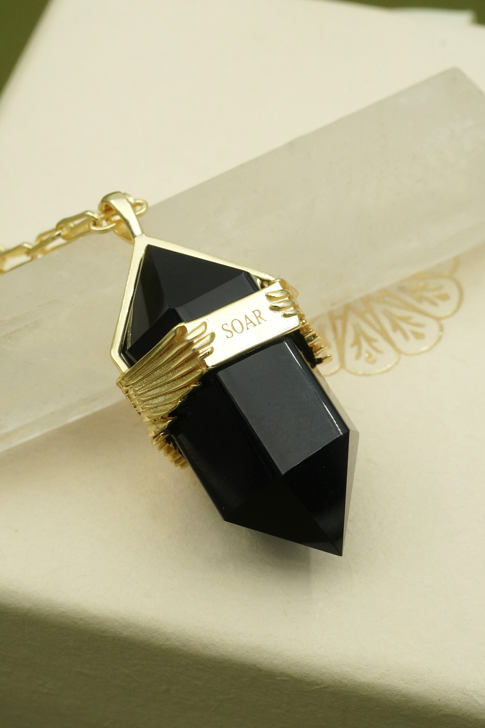 Gold spiritual necklace with black pendant inscribe wit the word soar for new beginnings on a beige background
