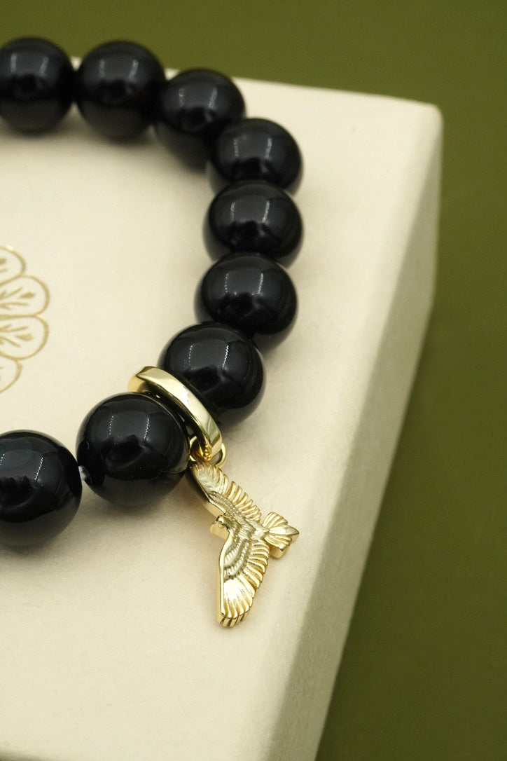 Black beaded gemstone bracelet with a gold soar bird charm on a white box against a green background