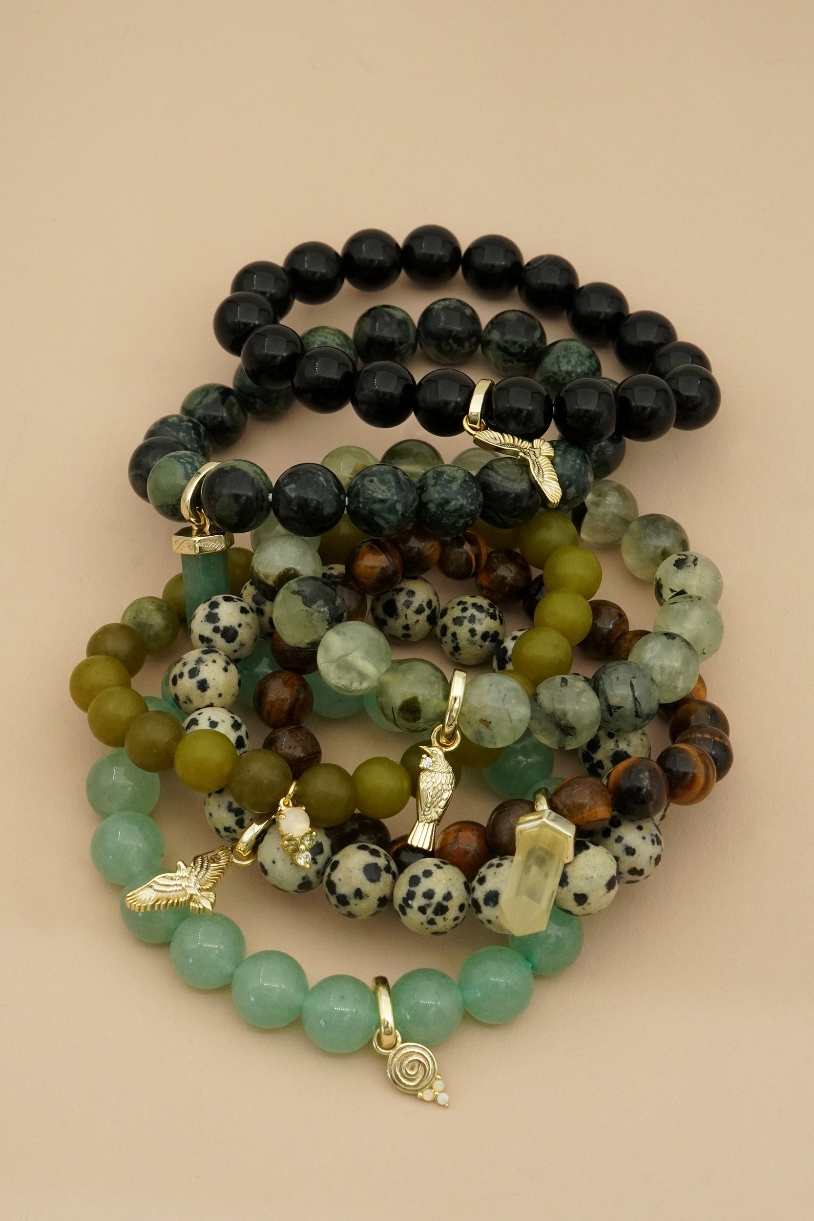 Stack of beaded gemstone bracelets with various earthy natural colours and gold charms on a beige background
