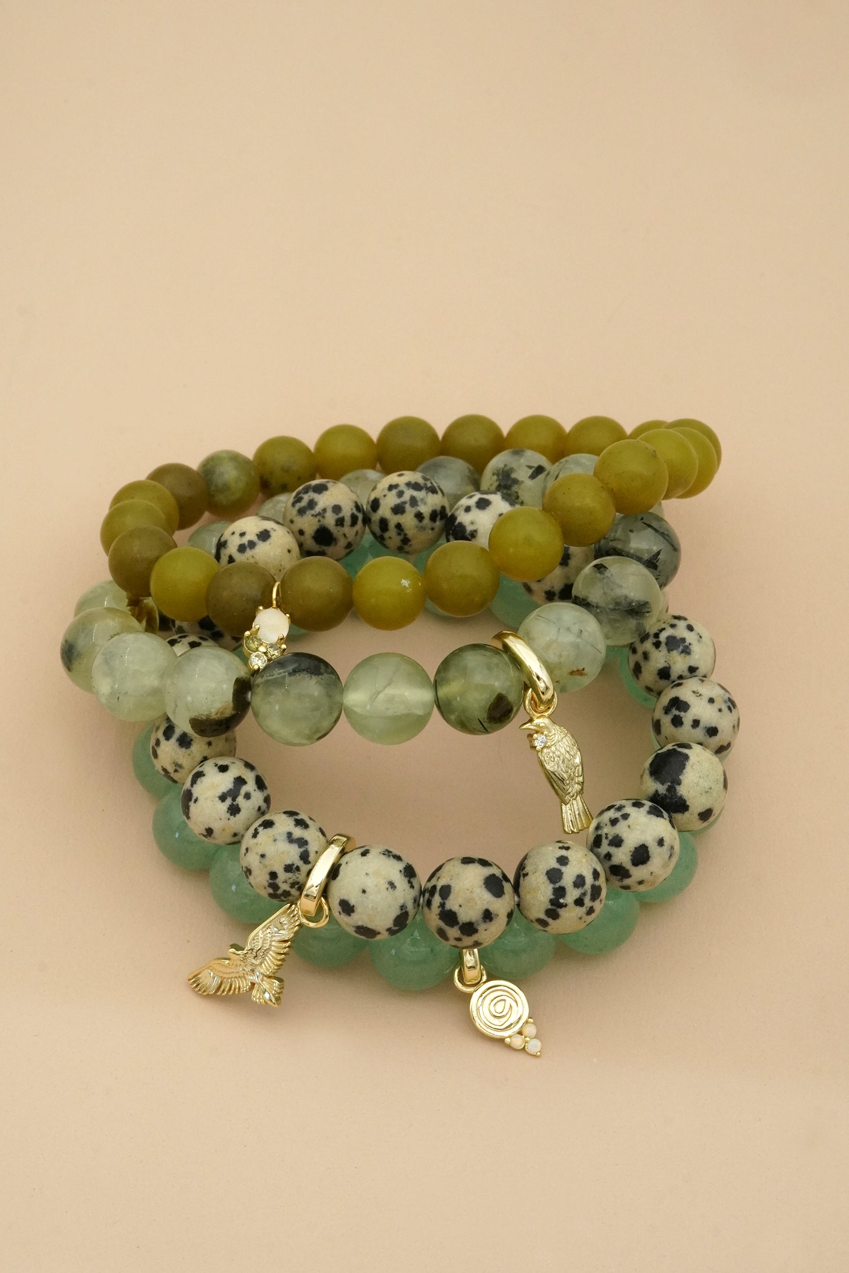 Stack of green and black beaded bracelets with gold bird charms on a beige background