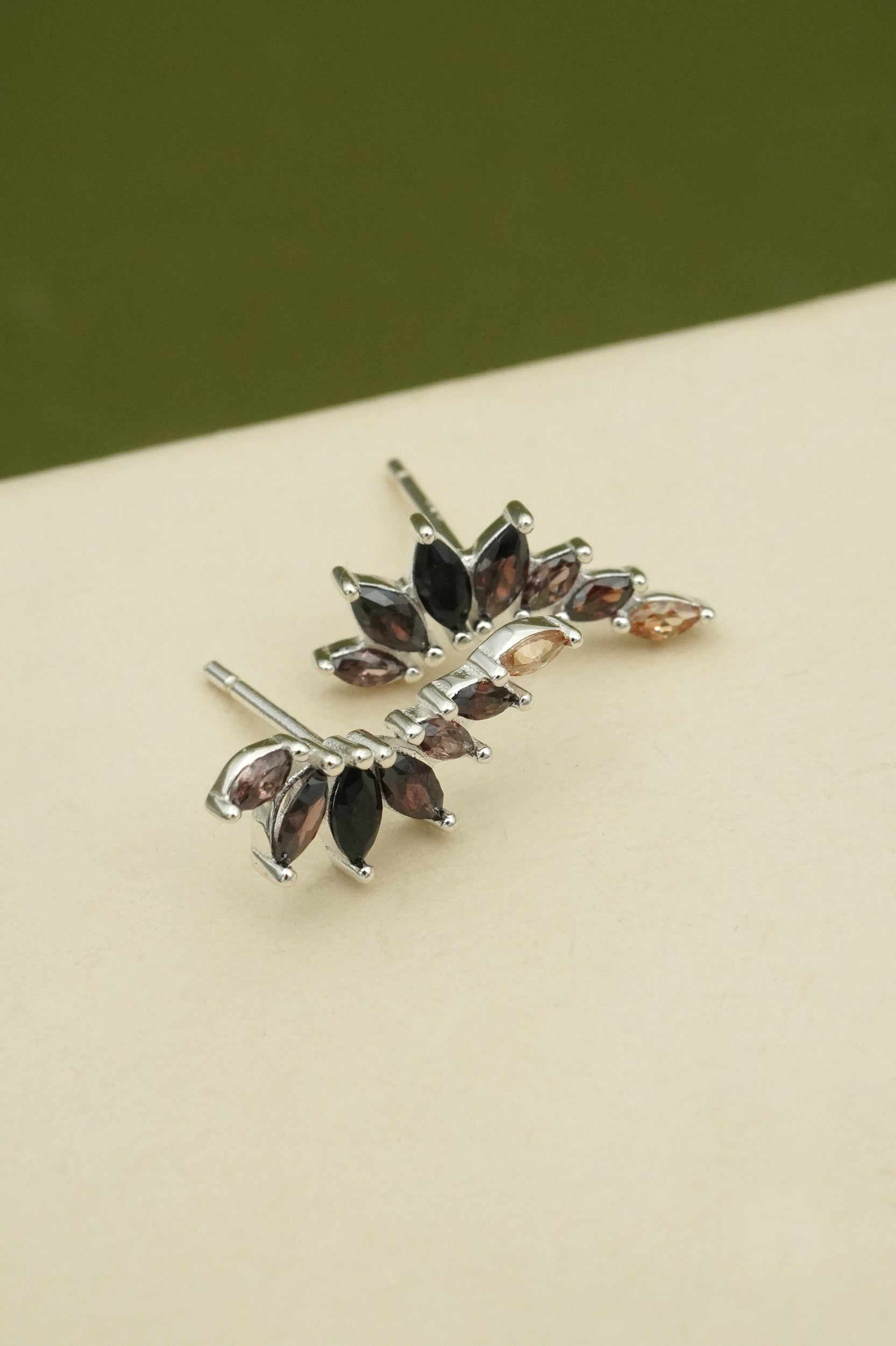 Silver leaf-shaped earrings with gemstones on a beige surface and green background