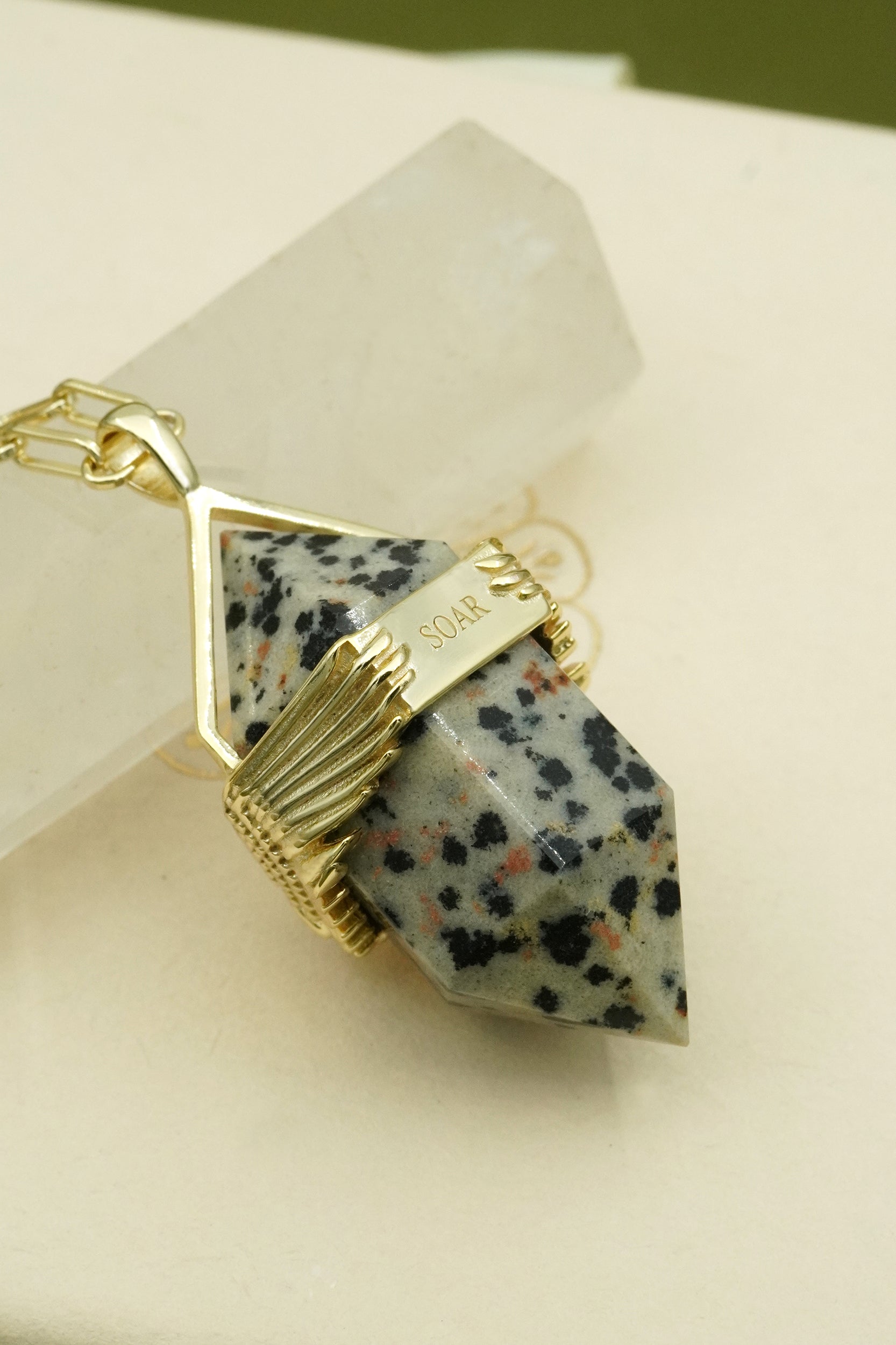 Gold Dalmatian jasper crystal pendant with words soar inscribed on a beige surface