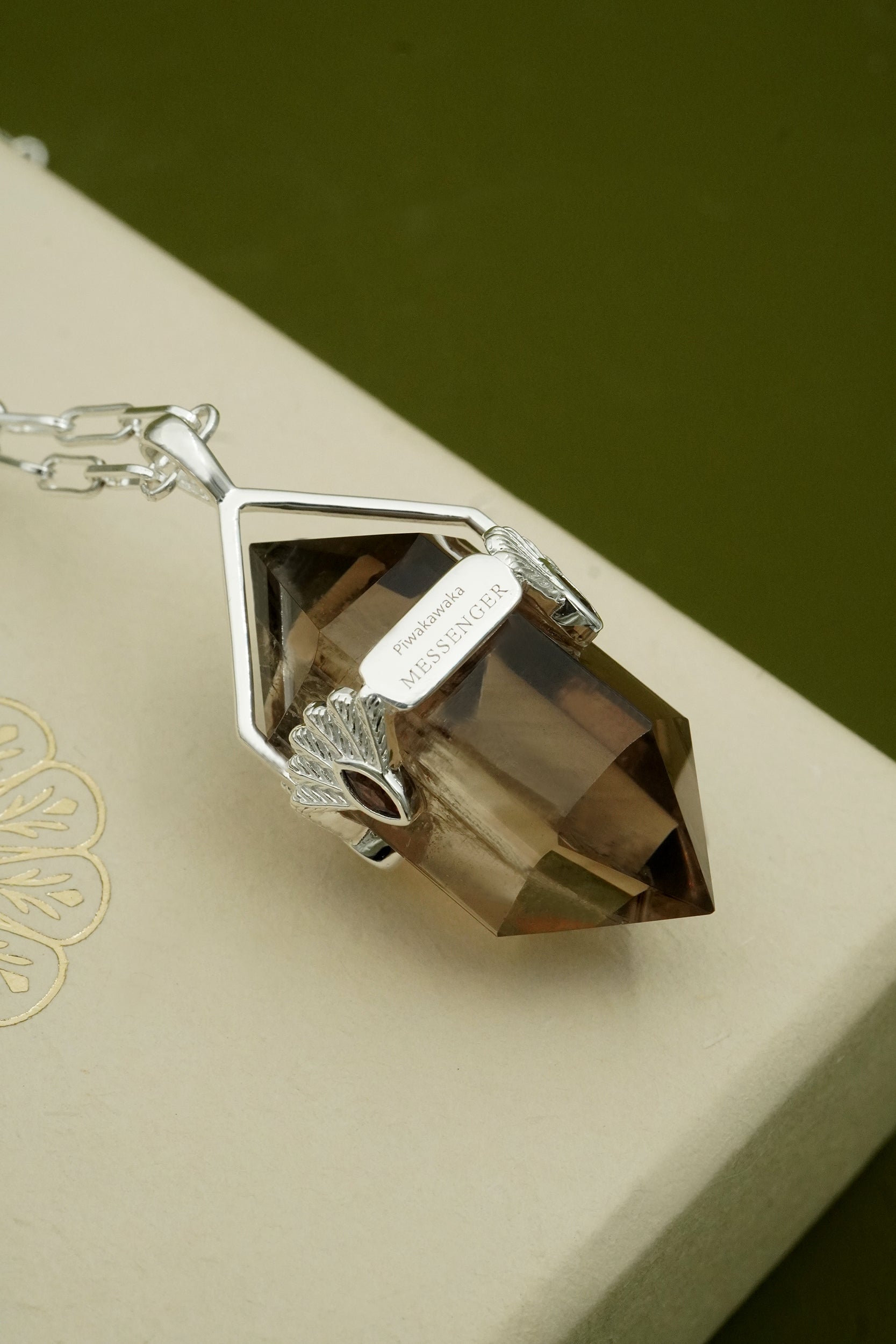 Silver necklace with a large brown gemstone on a beige surface. Inscription with words Piwakawaka Messenger.