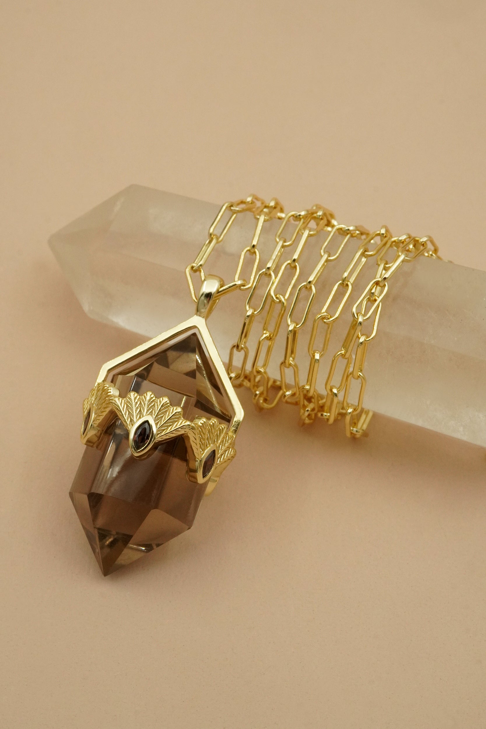 Gold piwakawaka necklace with a smoky crystal pendant on a beige background