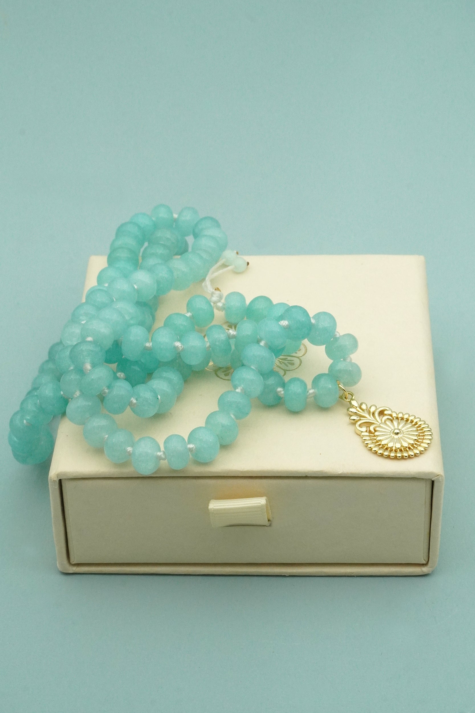 Turquoise beaded prayer male necklace with a gold charm on a beige box against a light blue background