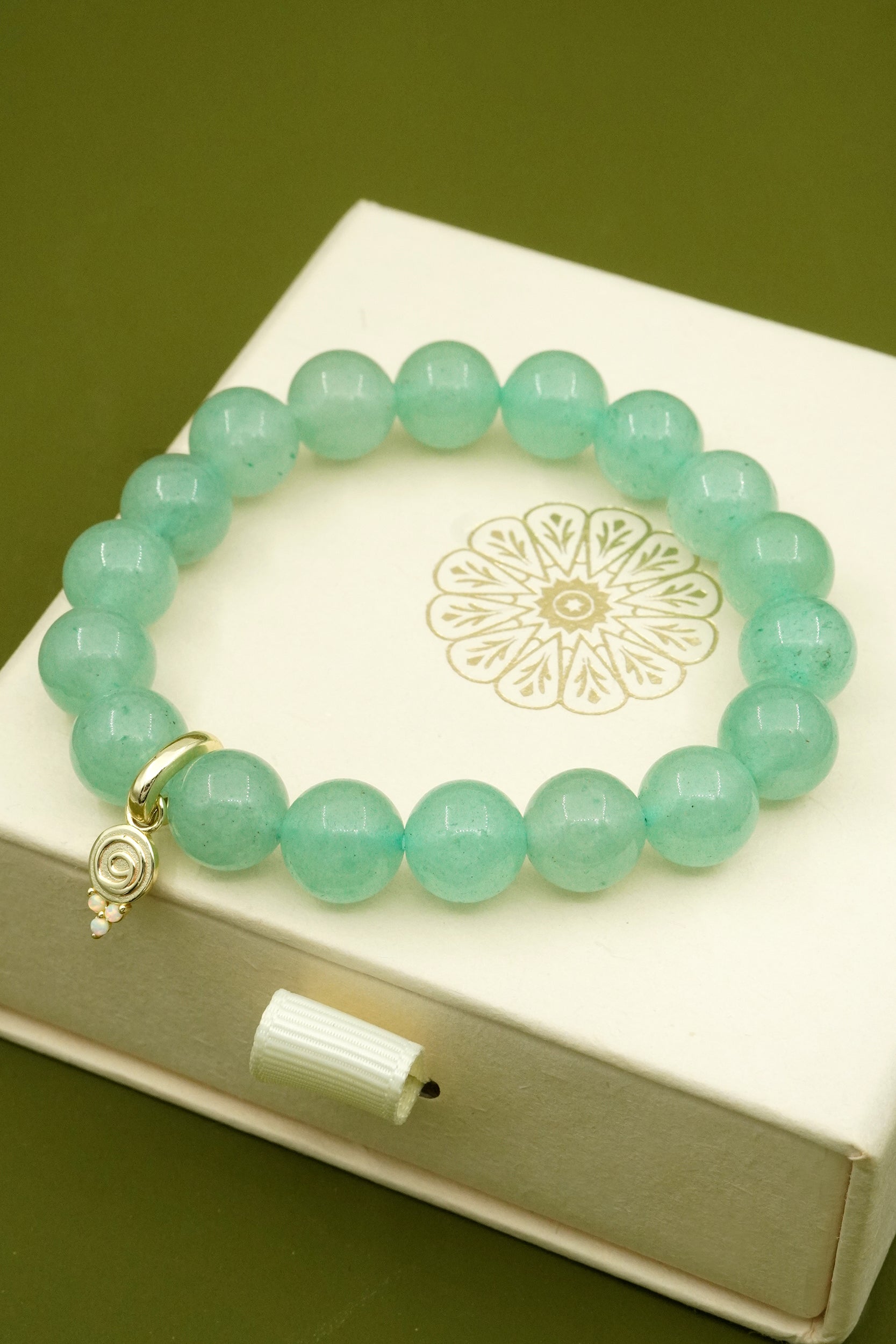 Green beaded crystal bracelet with a gold gratitude symbol charm on a white jewellery box with cloud nine jewellery logo.