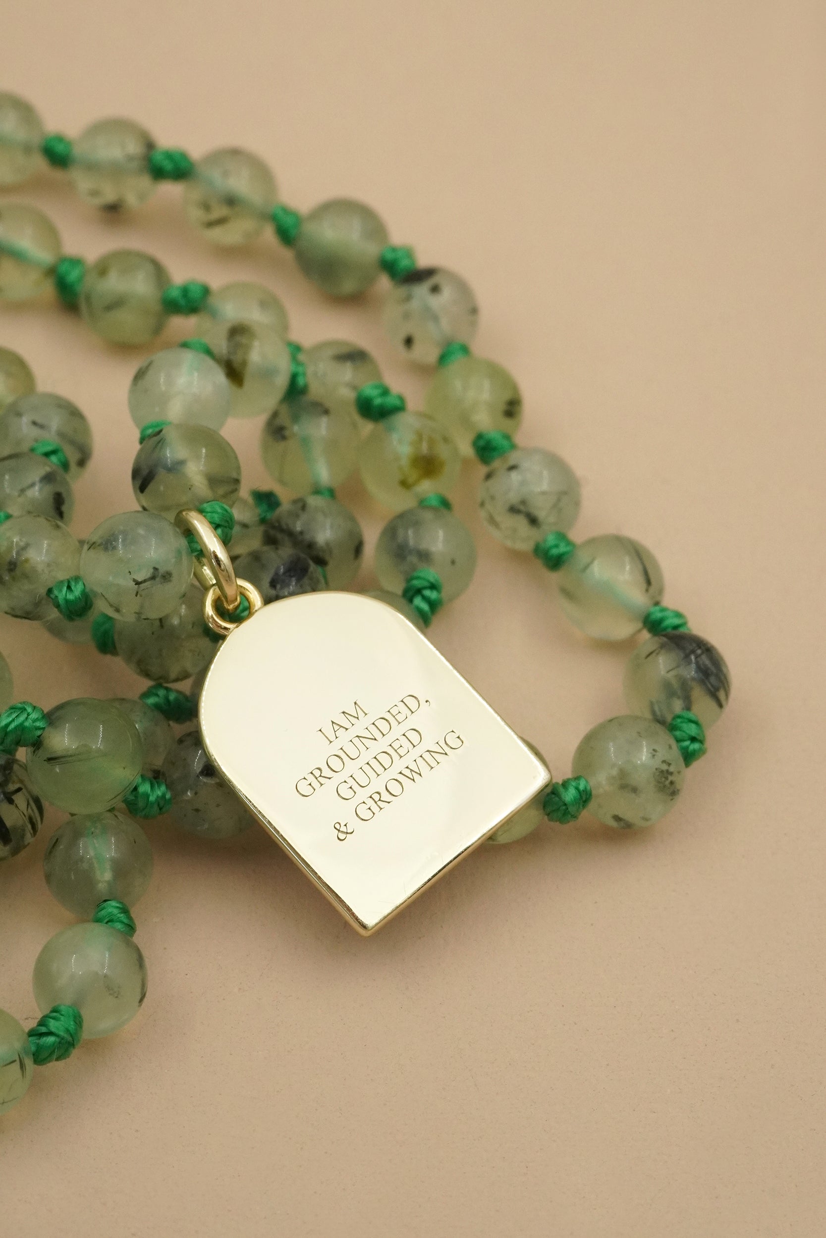 Mala Necklace with green beads and a gold pendant inscribed with I am grounded guided and growing on a beige background