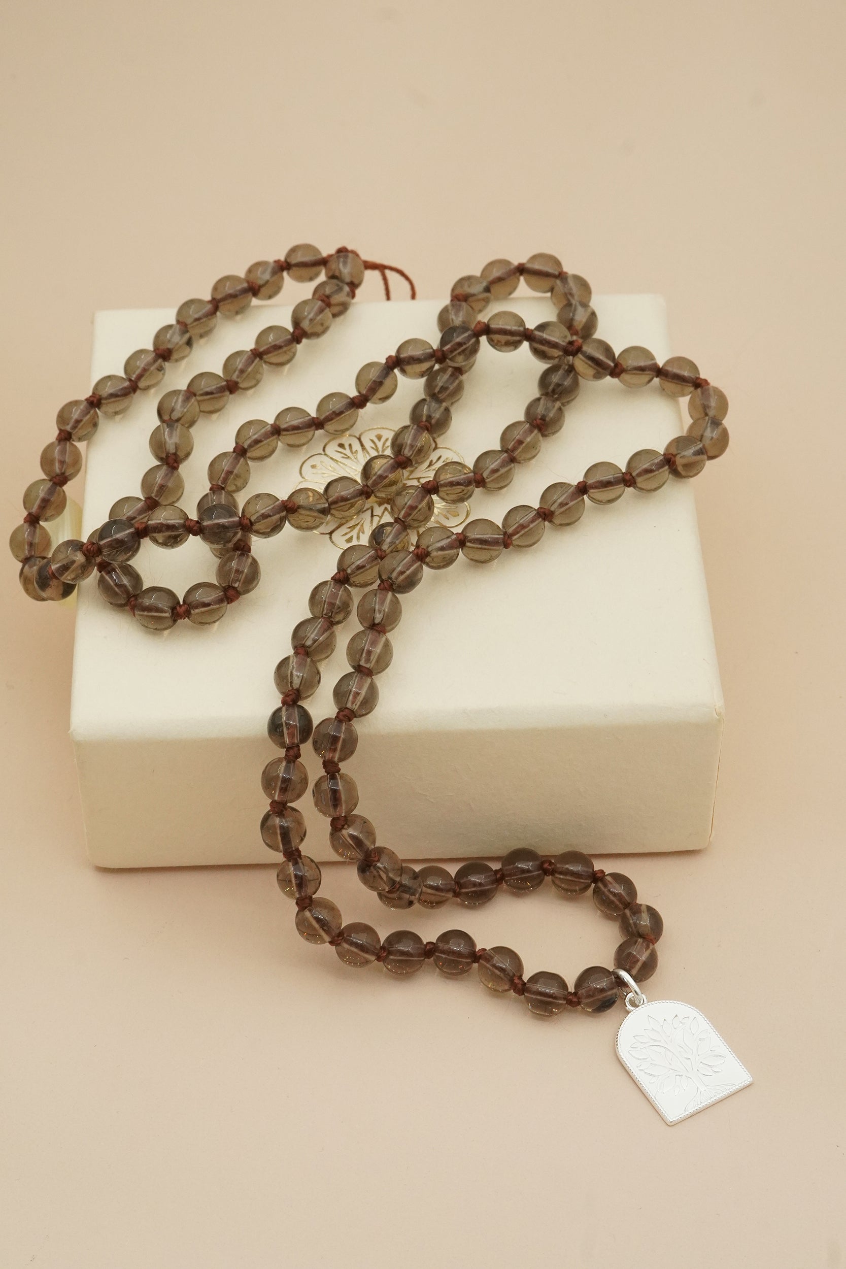 Brown beaded mala necklace with a silver tree of life pendant on a beige box against a beige background