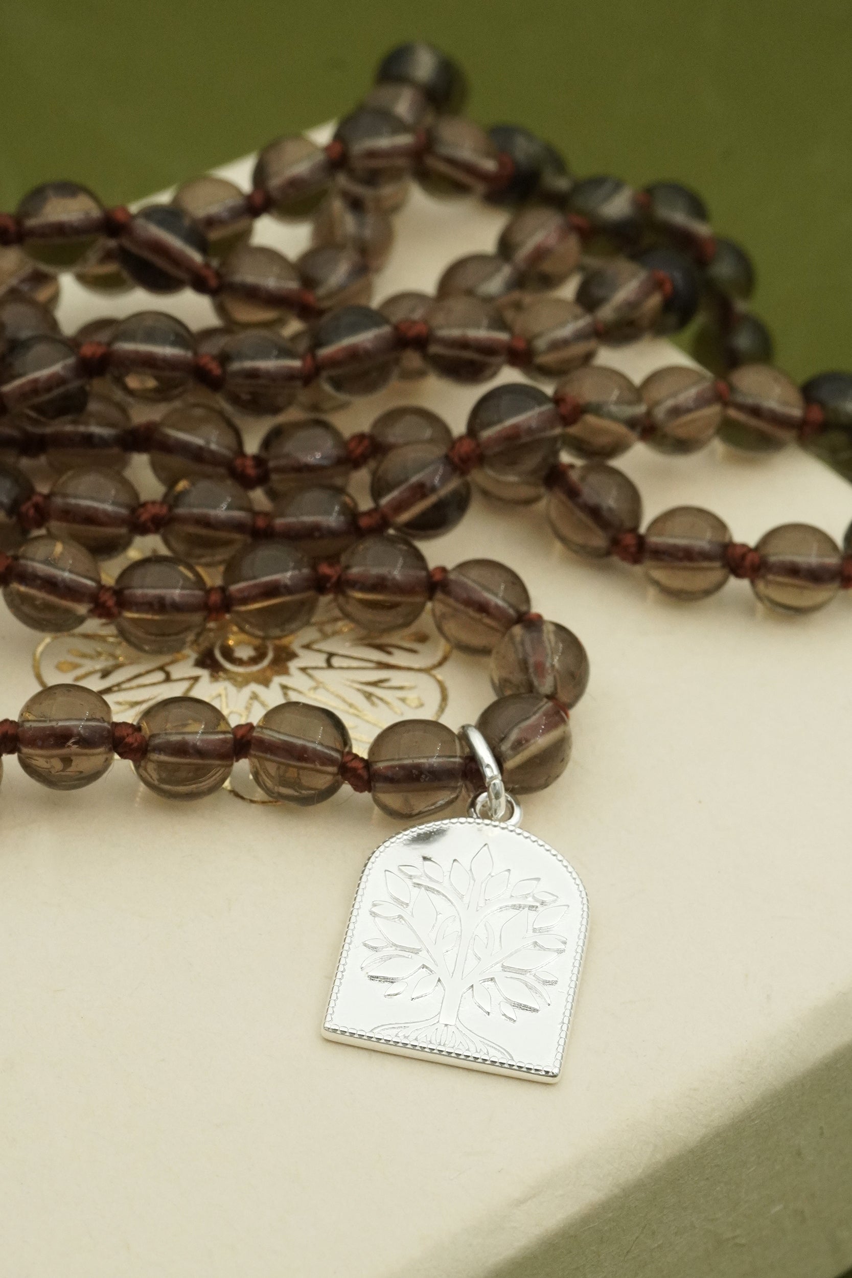Brown beaded mala necklace with a silver tree of life pendant on a beige surface