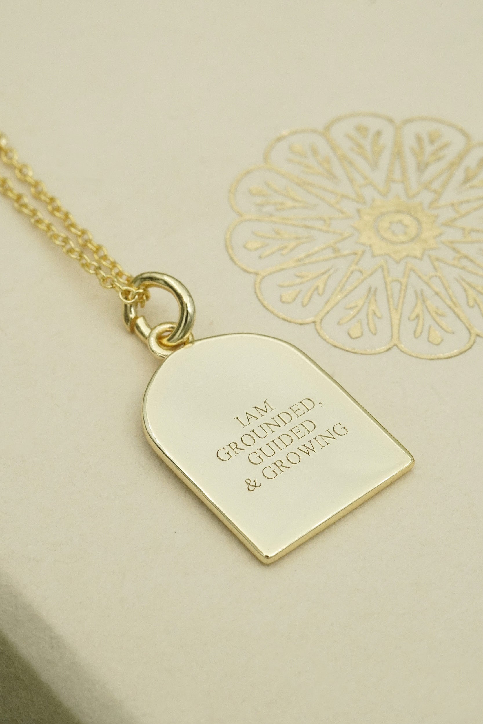 Gold necklace with a pendant on a beige background with inscription I am grounded, guided and growing