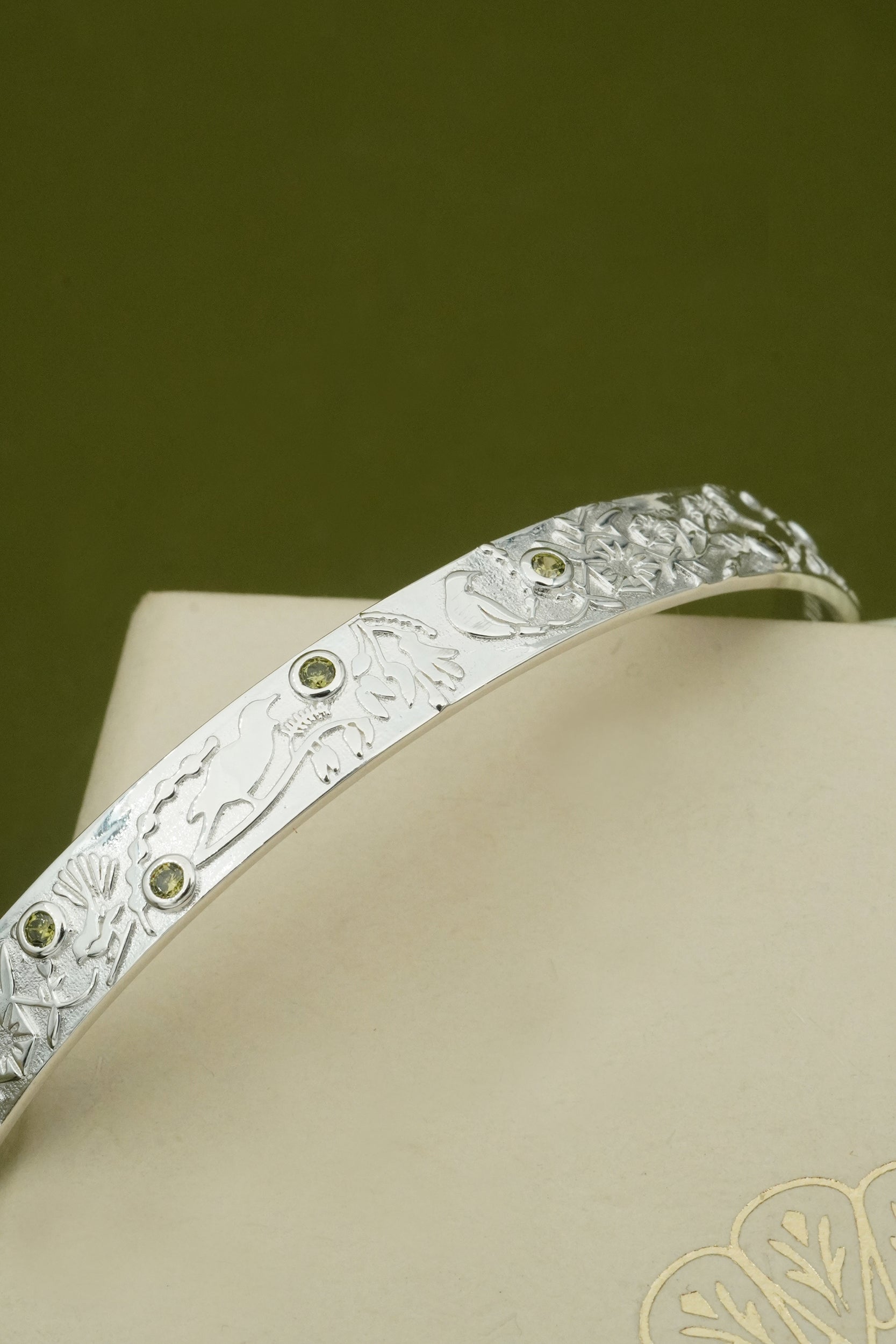 Silver cuff bracelet with engraved design and green gemstones on a beige surface with a green background