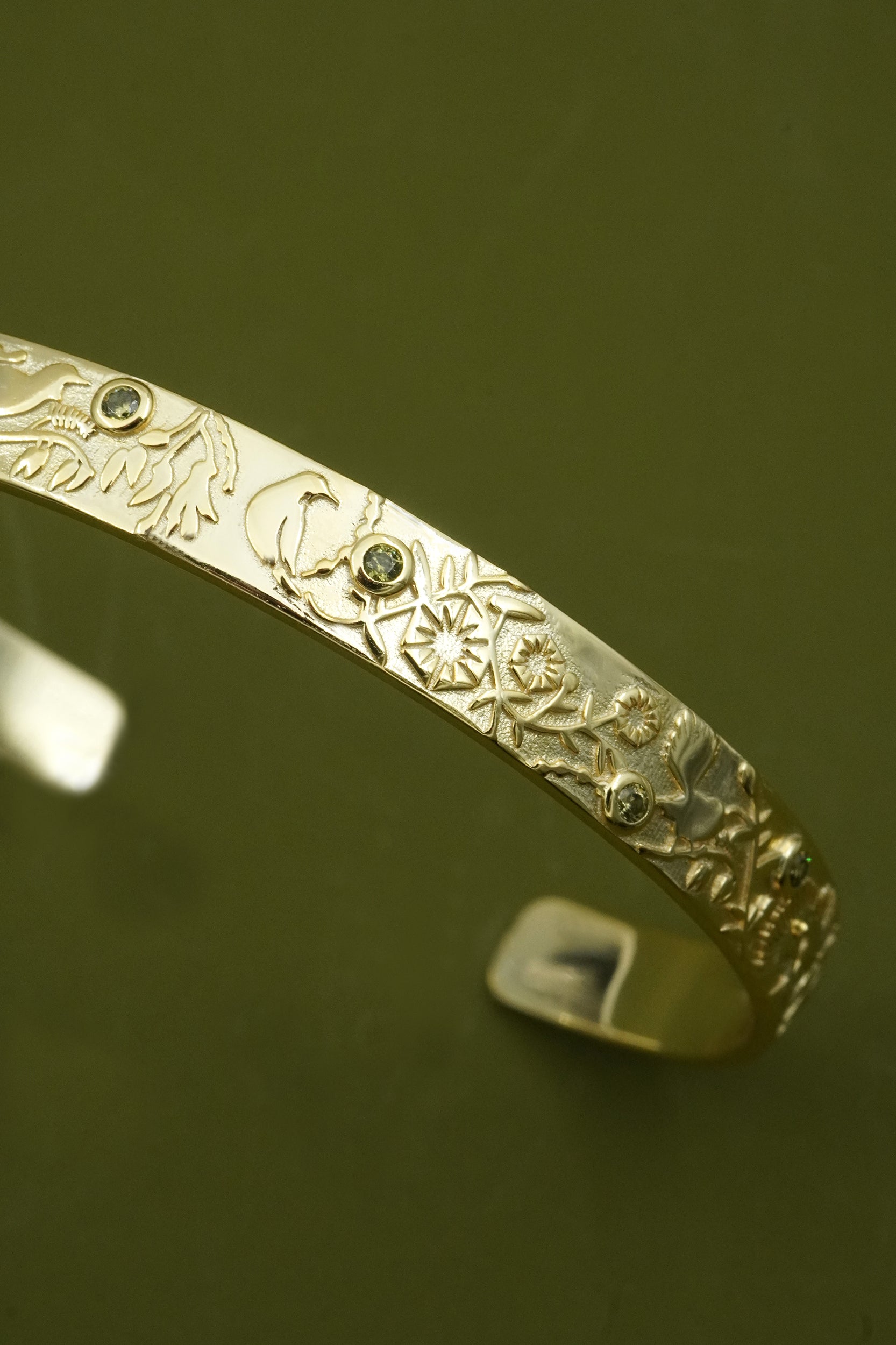 Gold cuff bracelet with intricate NZ forest designs and green gemstones on a reflective surface