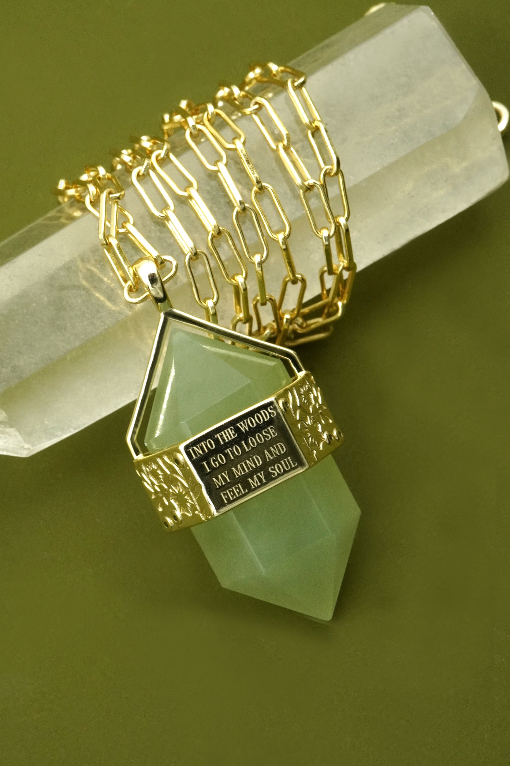 Gold necklace with a green stone pendant on a green background. Featuring the quote into the woods I go to loose my mind and feel my soul.