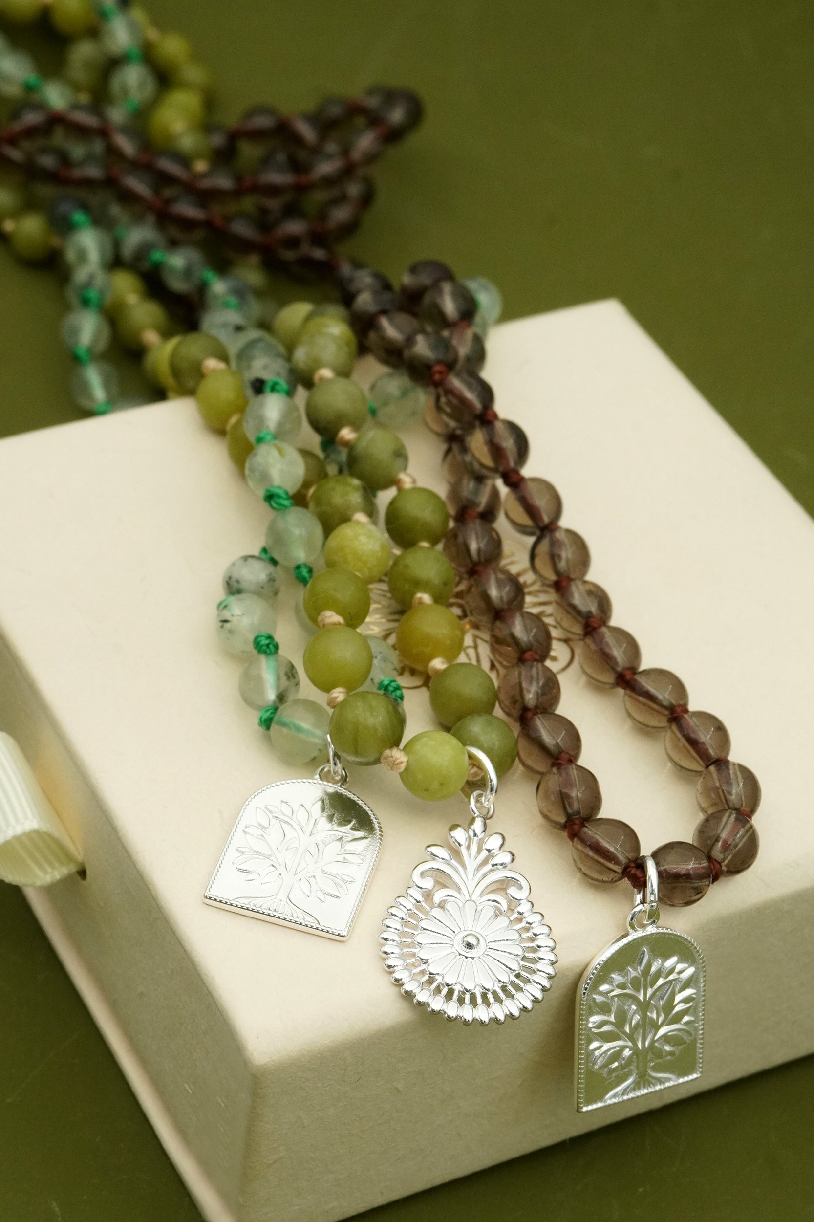 Three beaded spiritual male prayer necklaces with silver charms on a beige surface.