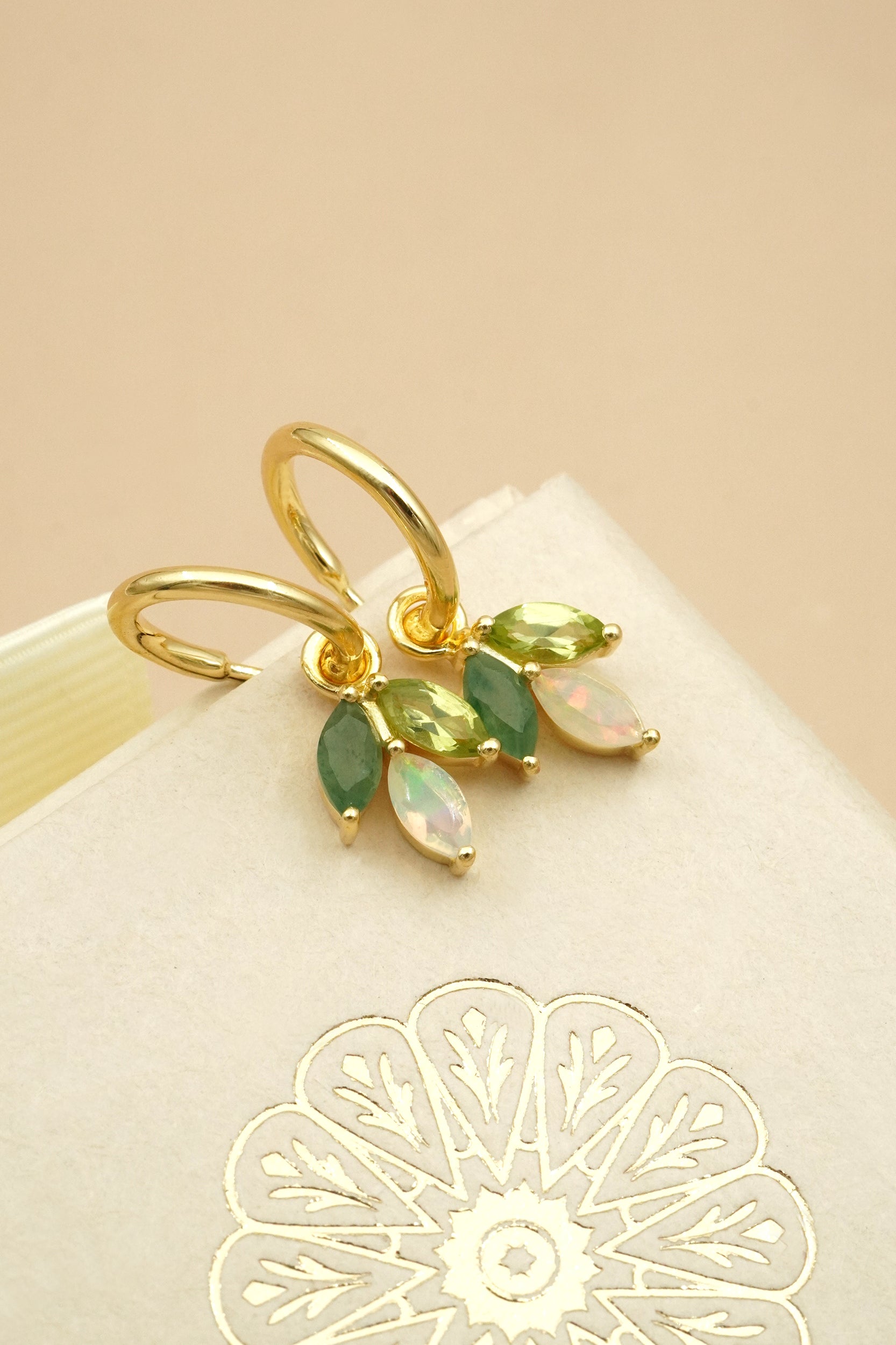 Gold floral earrings with green and opal stones on a beige background