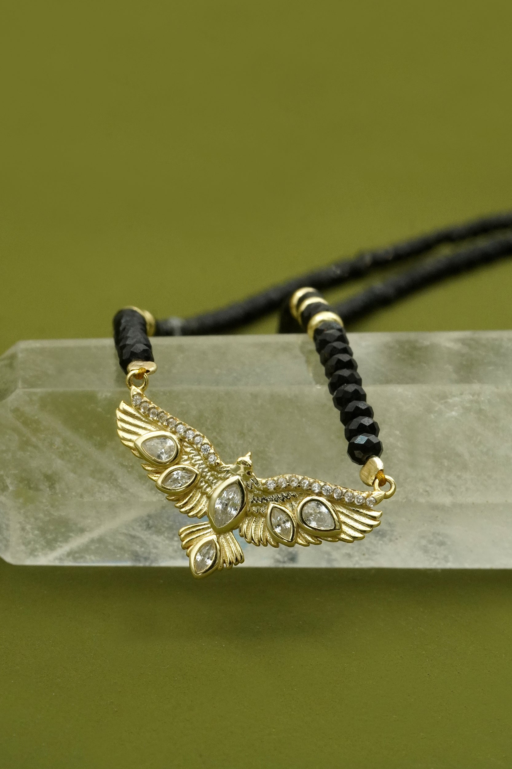 Gold eagle wings pendant on a black beaded chain against a green background