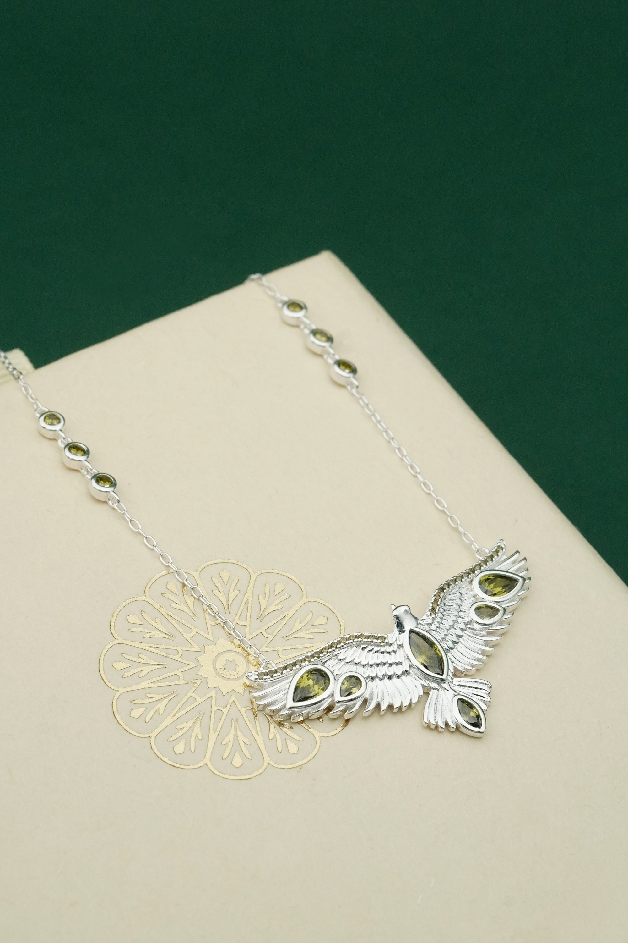 Silver bird pendant necklace with green stones on a beige surface