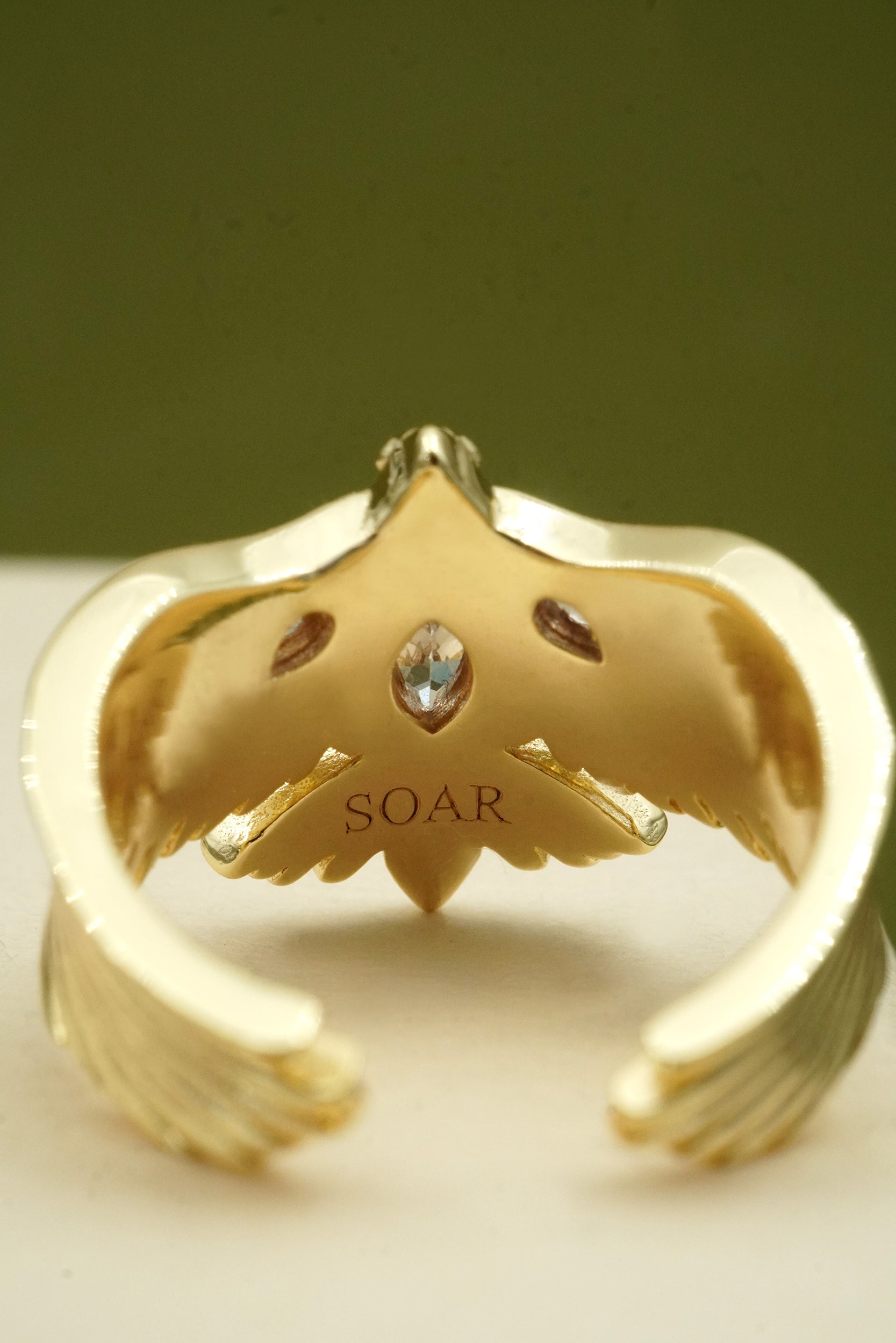 Gold bird-shaped ring with 'SOAR' engraving on a green background