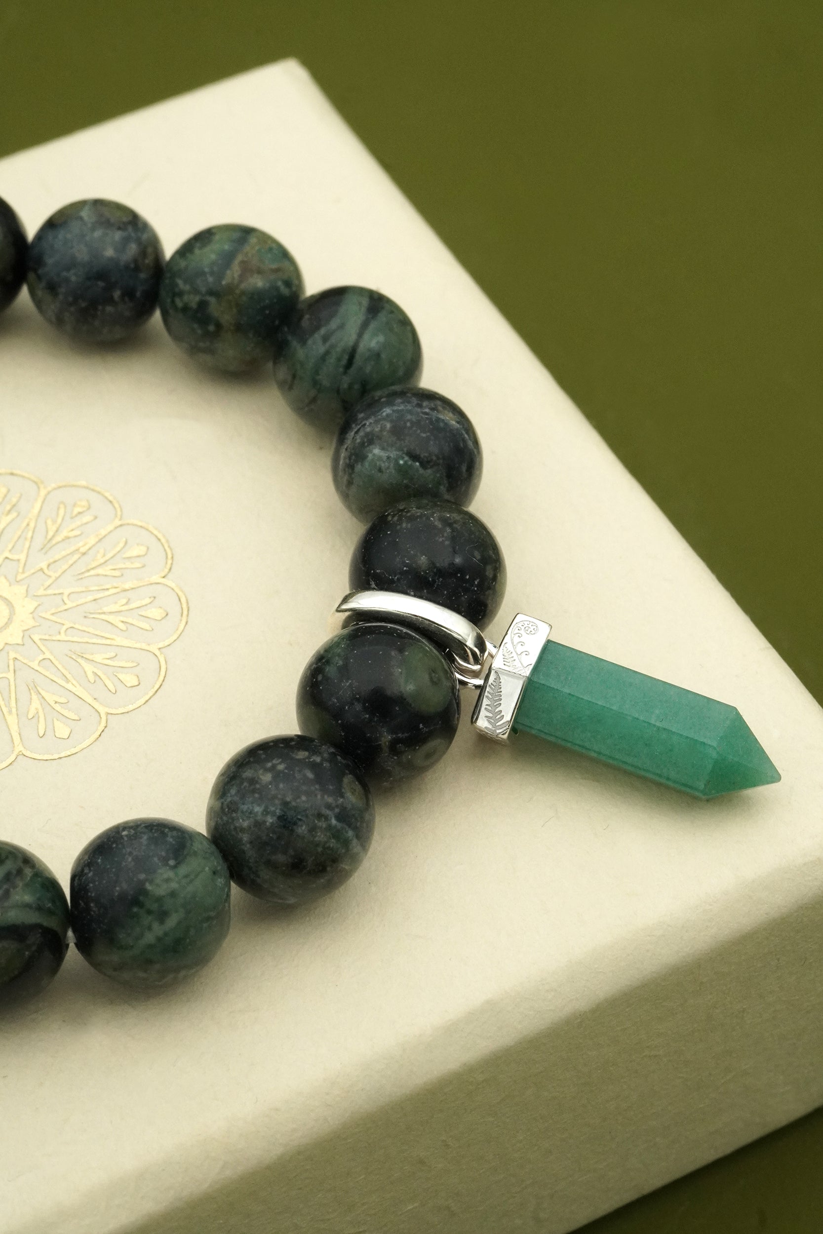 Beaded bracelet with a green crystal gemstones and a fern pendant on a white box with gold design