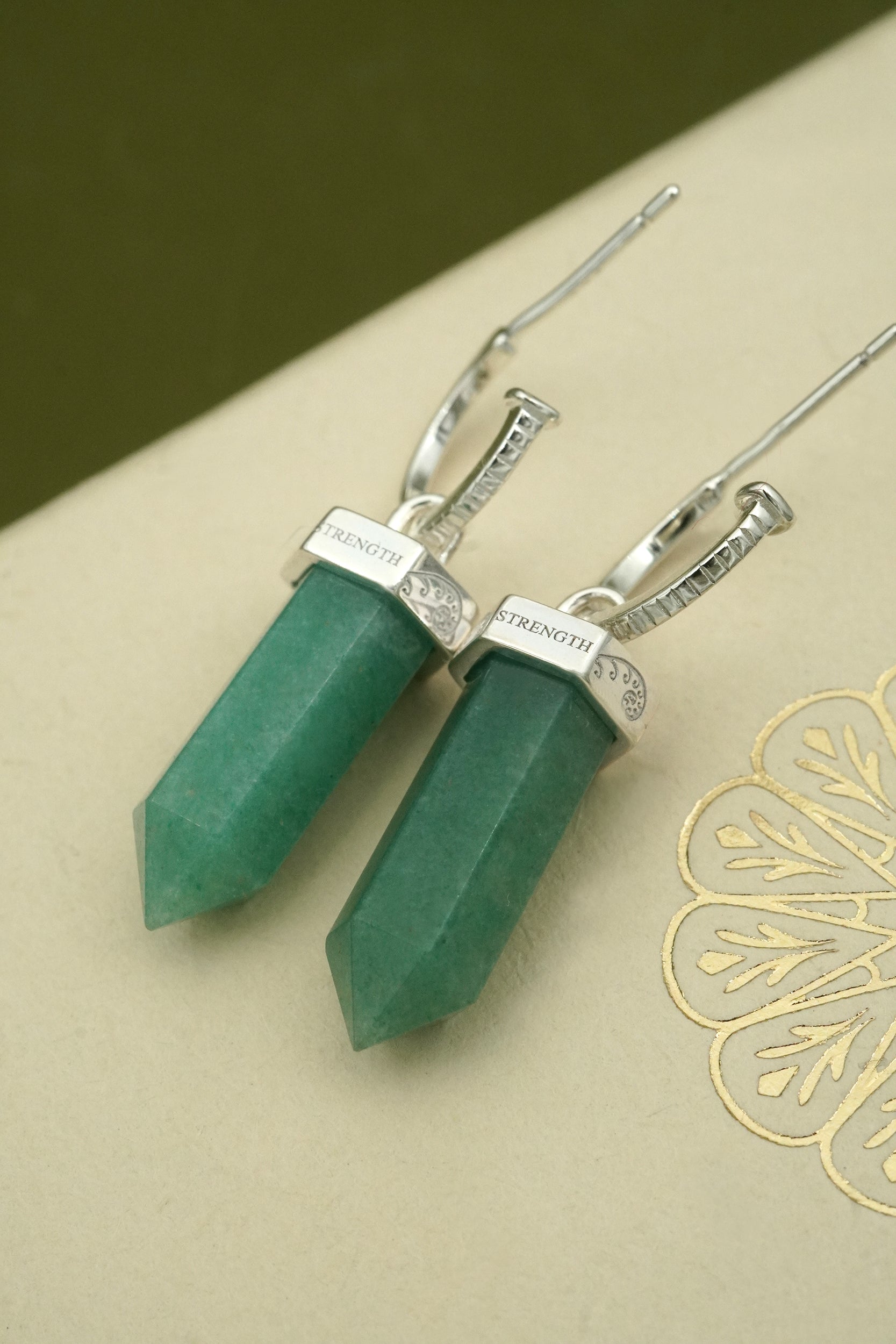 Green crystal pendant earrings on a beige surface with strength inscribed on the reverse