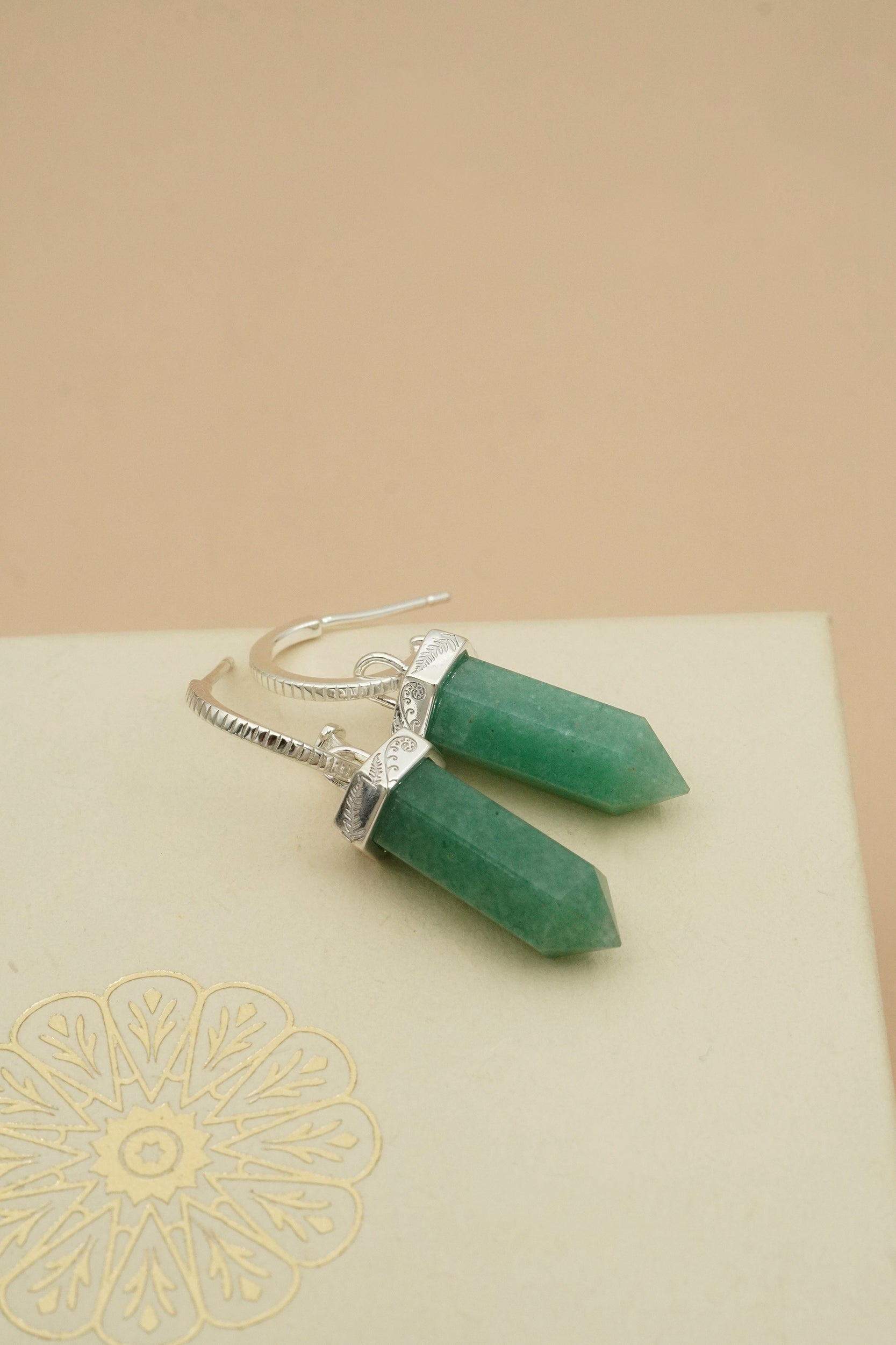 Green crystal earrings on a beige surface with a decorative Silver Fern and koru nature pattern