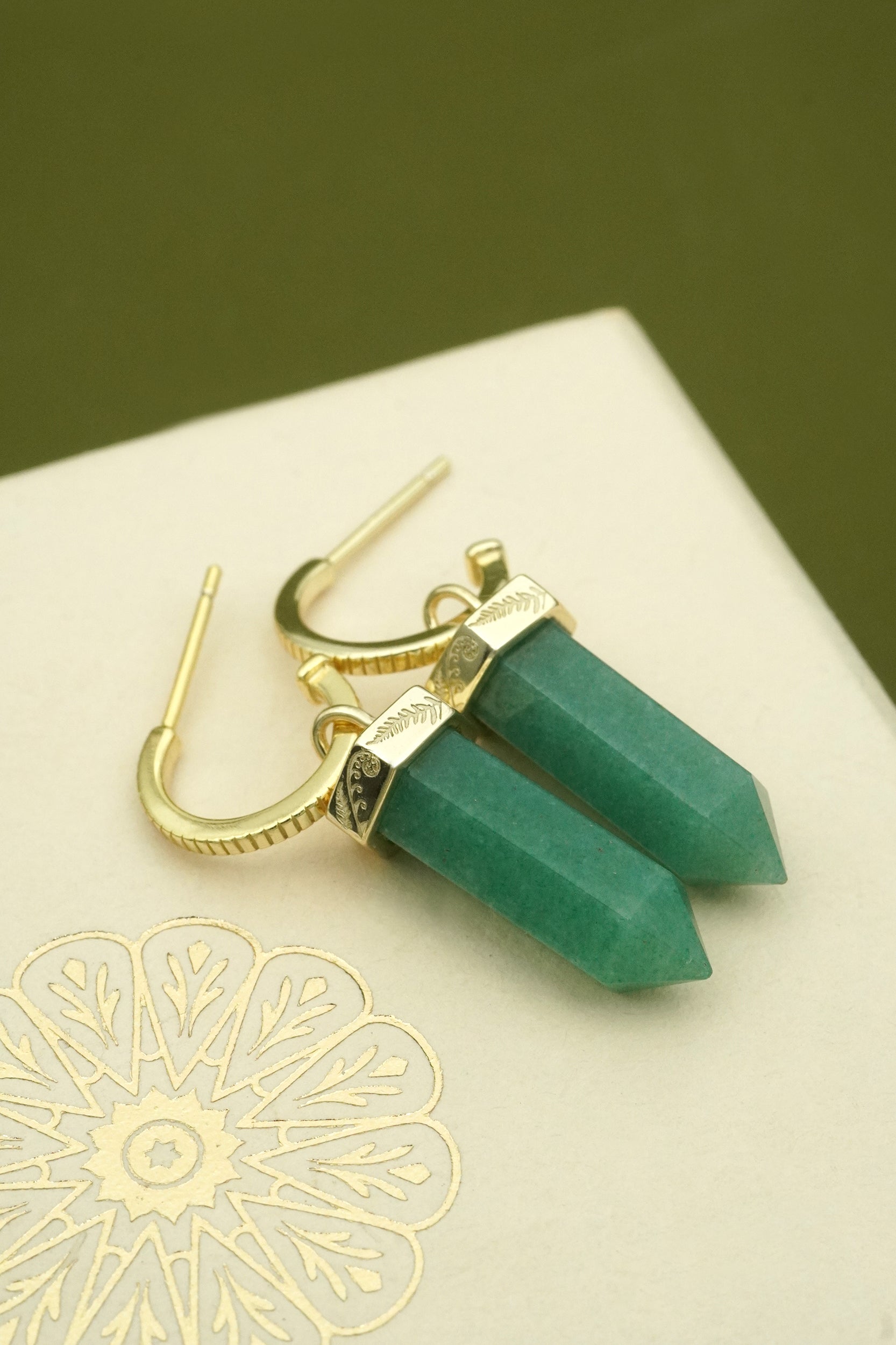 Green aventurine crystal earrings with koru and fern detailing on a decorative cream box