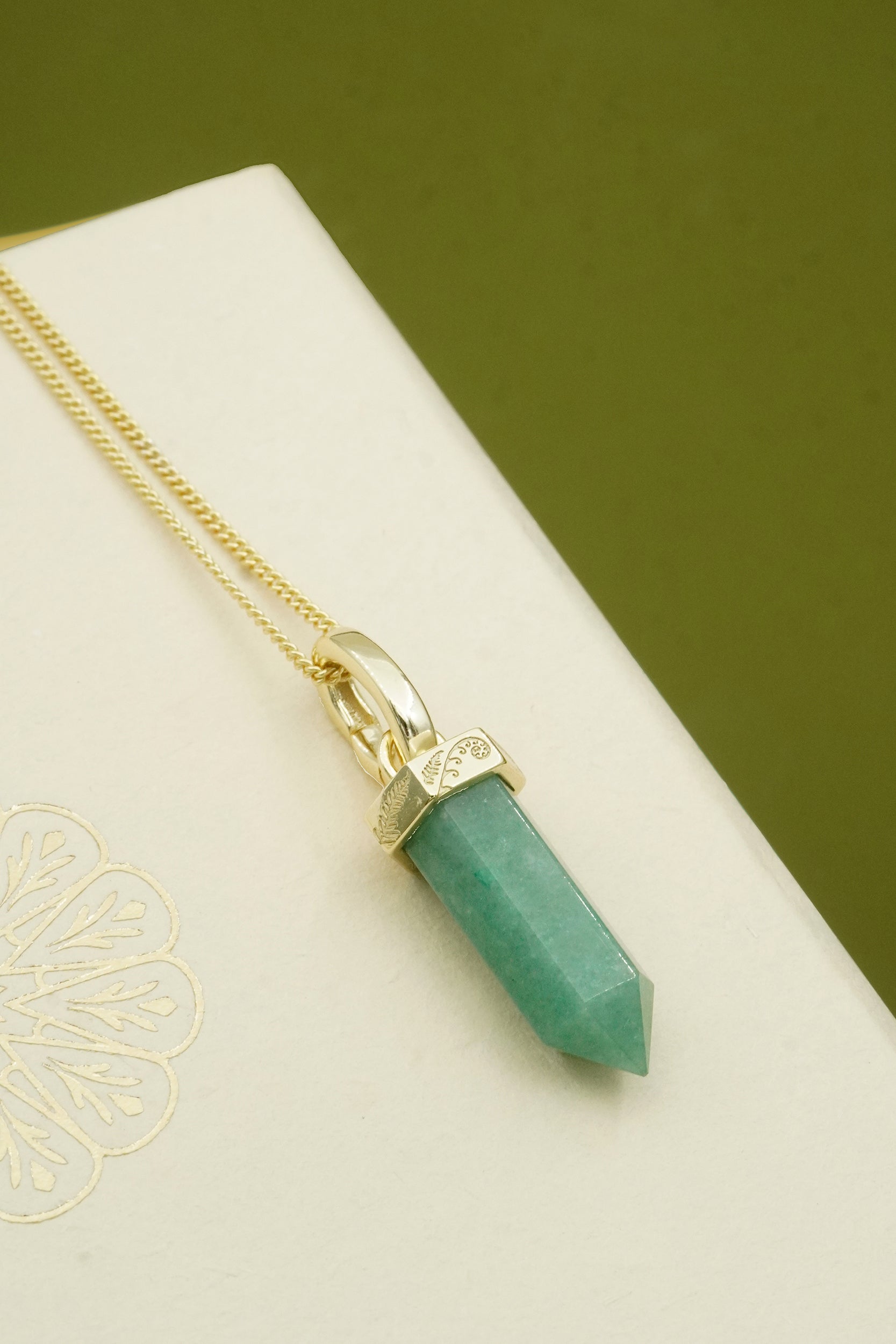 Green crystal pendant necklace on a gold chain featuring fern and koru detailing against a green background