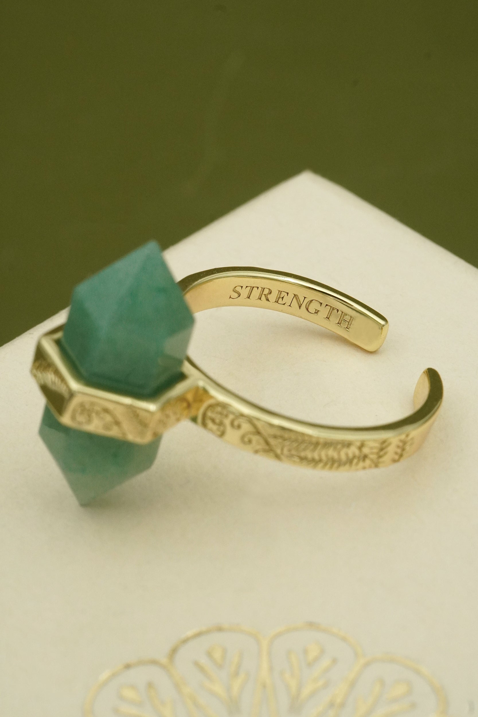 Gold bracelet with green gemstone and words strength engraved on inside of band on a white surface