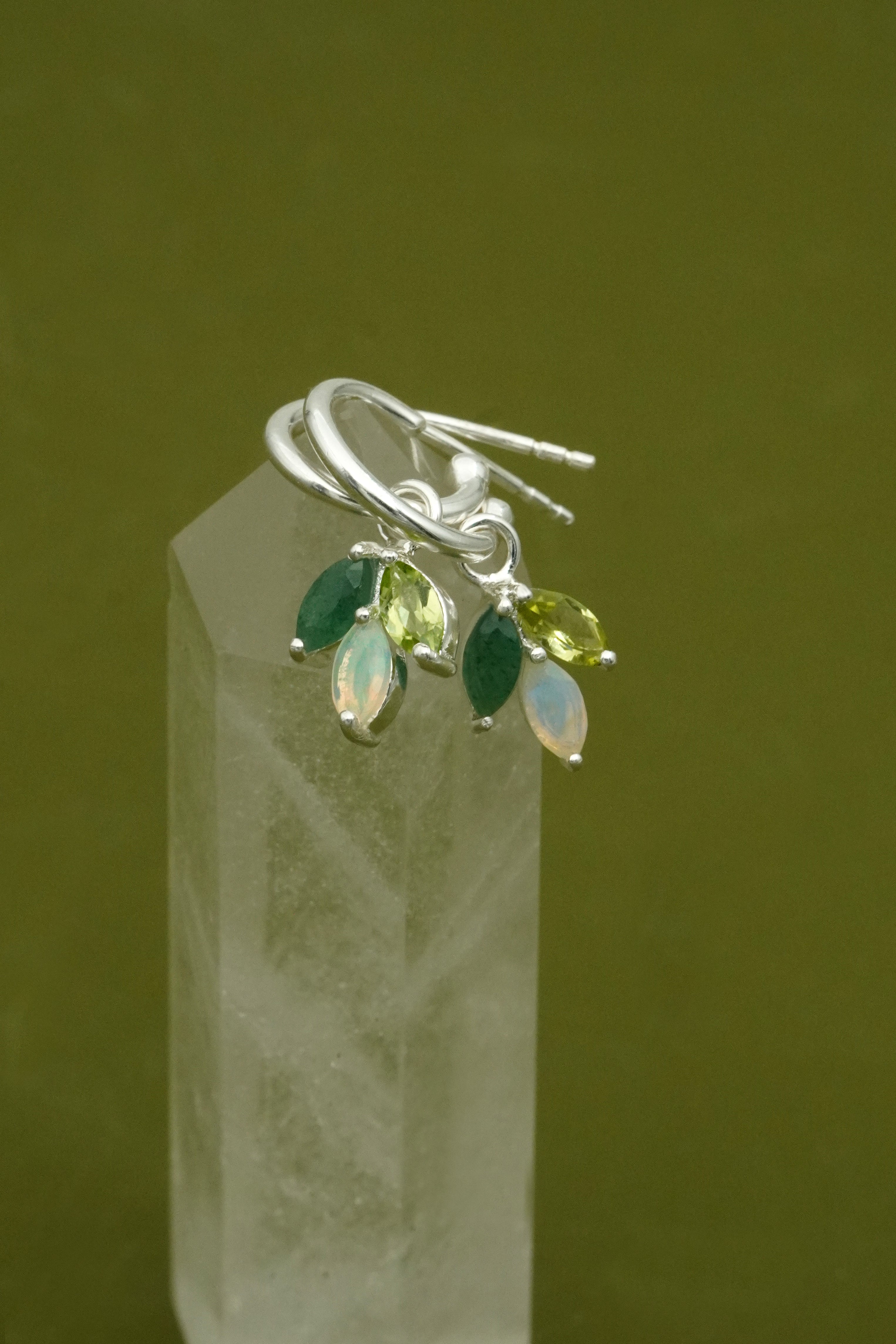 Silver earrings with green and clear gemstones on a clear crystal block against a green background
